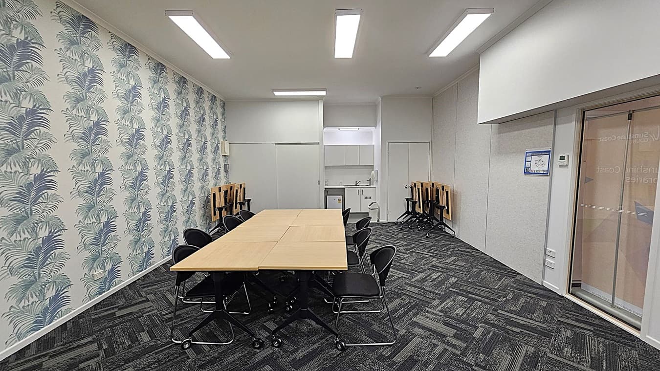 A meeting room with a long table in the centre surrounded by black chairs. On the left, the wall is decorated with a pattern of large leaves. The right side features a glass door and a plain wall with a poster. In the background, there is a small kitchenette with a sink and cabinets. The floor is covered with dark patterned carpet tiles, and the ceiling has rectangular fluorescent lights.