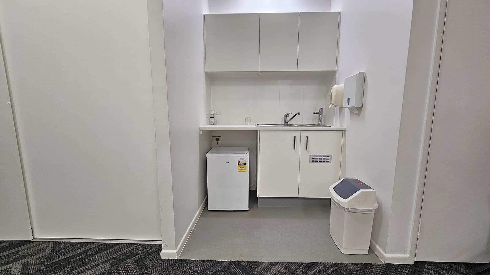 A small kitchenette with white cabinetry. On the left, there's a compact white fridge under the counter. In the centre, a sink with a tap is set into the countertop, with a soap dispenser and a glass nearby. Above, there are wall-mounted cabinets. On the right, a paper towel dispenser is mounted on the wall, and a white bin with a black lid is on the floor. The flooring is grey, and the walls are white.