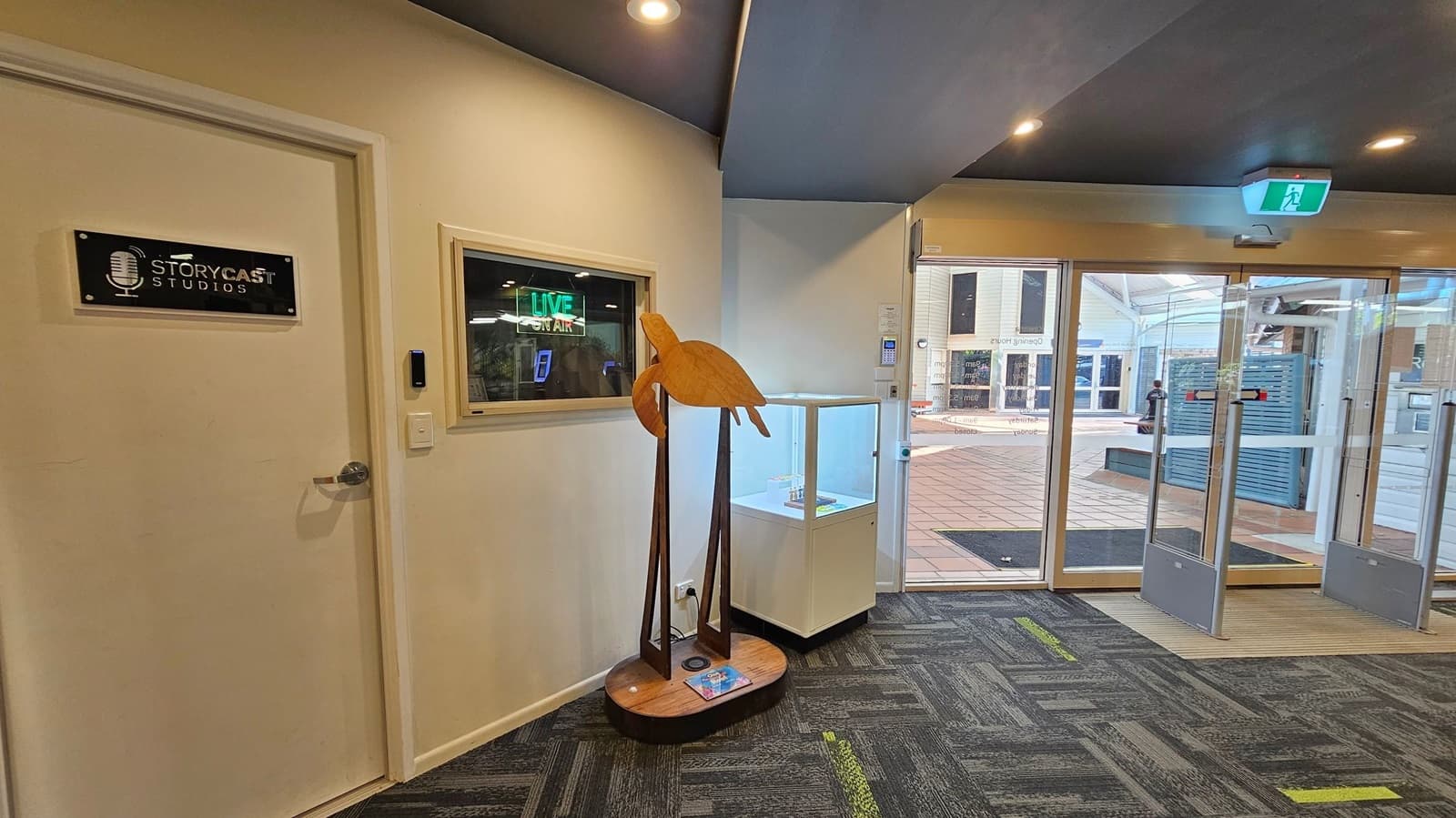A hallway with a door on the left labelled "StoryCast Studios" and a window displaying a "LIVE ON AIR" sign. In the centre, a wooden sculpture of a bird stands on a pedestal. To the right, a glass display case is positioned near a set of glass doors leading outside. The floor is covered with a patterned carpet in shades of grey and green, and the ceiling is dark with recessed lighting. An emergency exit sign is above the doors.