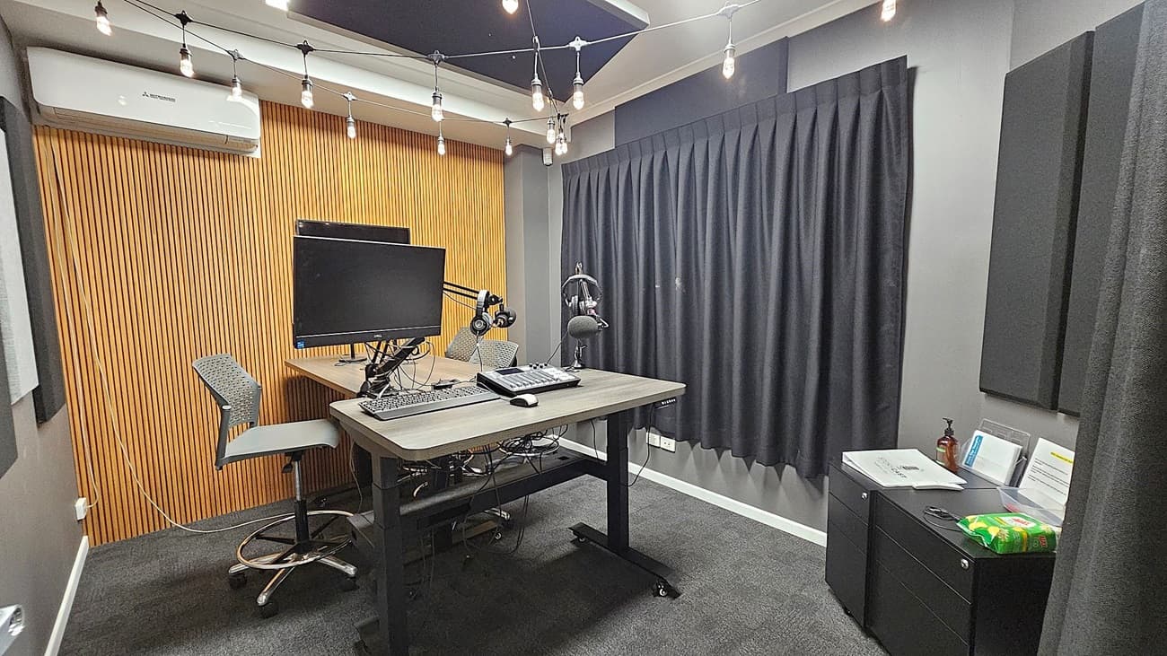 A modern recording studio with a wooden slat wall on the left and a grey curtain on the right. In the centre, there's a desk with a computer monitor, keyboard, headphones, and a microphone on an adjustable arm. Two high chairs are positioned at the desk. The ceiling has string lights hanging across. On the right, a small cabinet holds papers, a bottle, and a packet of snacks. The floor is carpeted in dark grey.