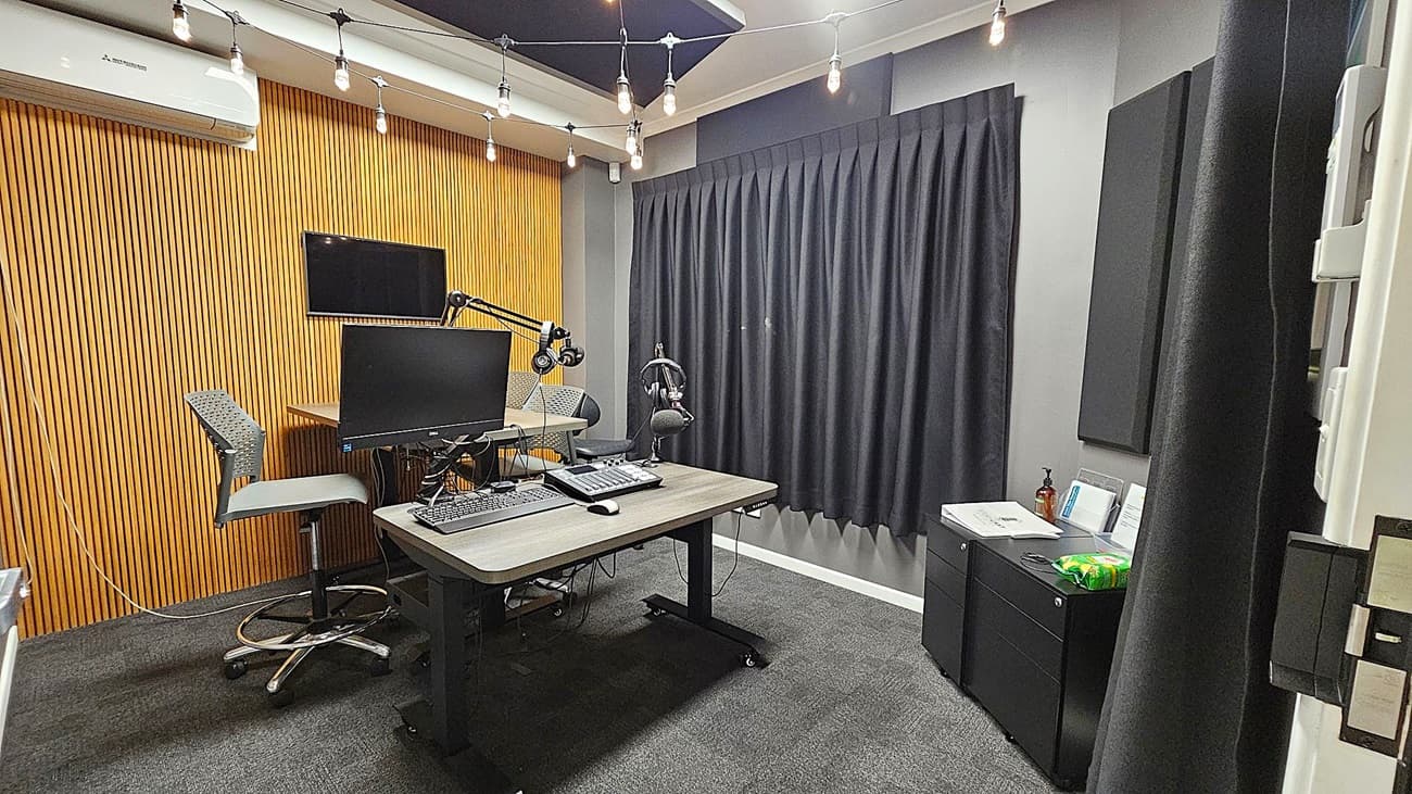 A recording studio with a wooden panelled wall on the left, featuring an air conditioning unit and a mounted monitor. Two grey chairs are positioned at a desk with a computer monitor, keyboard, and microphone. The right side has a black curtain and a small cabinet with papers and a bottle on top. The ceiling has exposed light bulbs, and the floor is carpeted in dark grey.