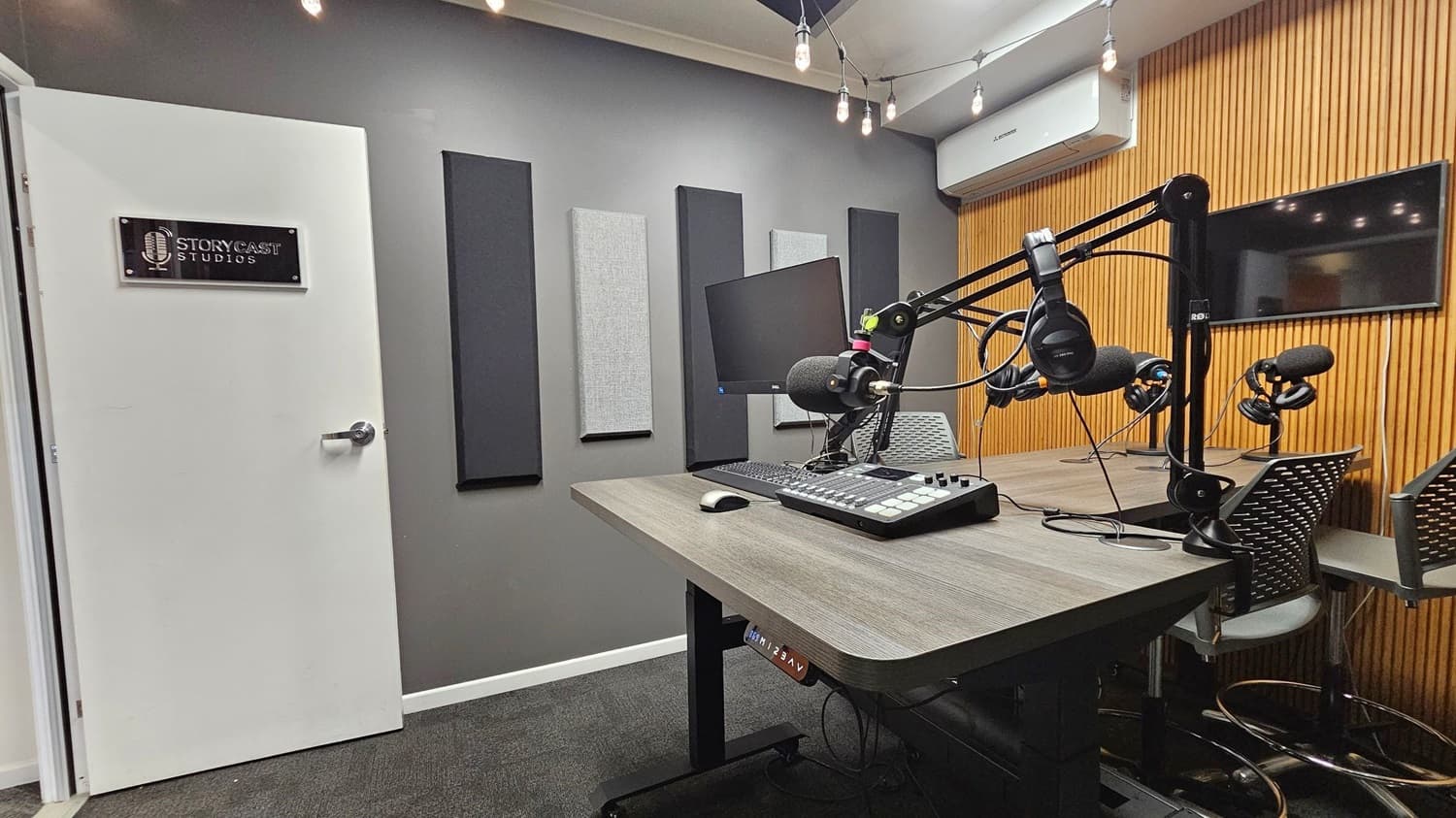 A podcast studio with a white door on the left featuring a "Storycast Studios" sign. The grey wall has acoustic panels. In the centre, a wooden table holds a computer monitor, keyboard, and audio mixer. Microphones and headphones are mounted on adjustable arms. The right wall is lined with vertical wooden slats and a flat-screen TV. The ceiling has exposed light bulbs, and the floor is carpeted in dark grey.