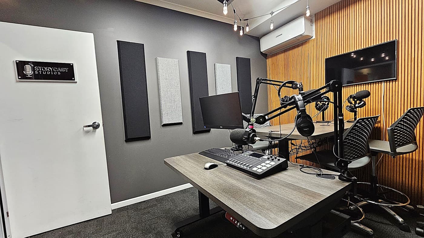 A podcast studio with a grey wall on the left featuring acoustic panels and a white door with a "Storycast Studios" sign. In the centre, there's a desk with a microphone, headphones, a computer monitor, and audio equipment. On the right, a wooden slat wall with a mounted screen and two chairs. The ceiling has hanging light bulbs, and the floor is carpeted in dark grey. An air conditioning unit is mounted above the wooden wall.