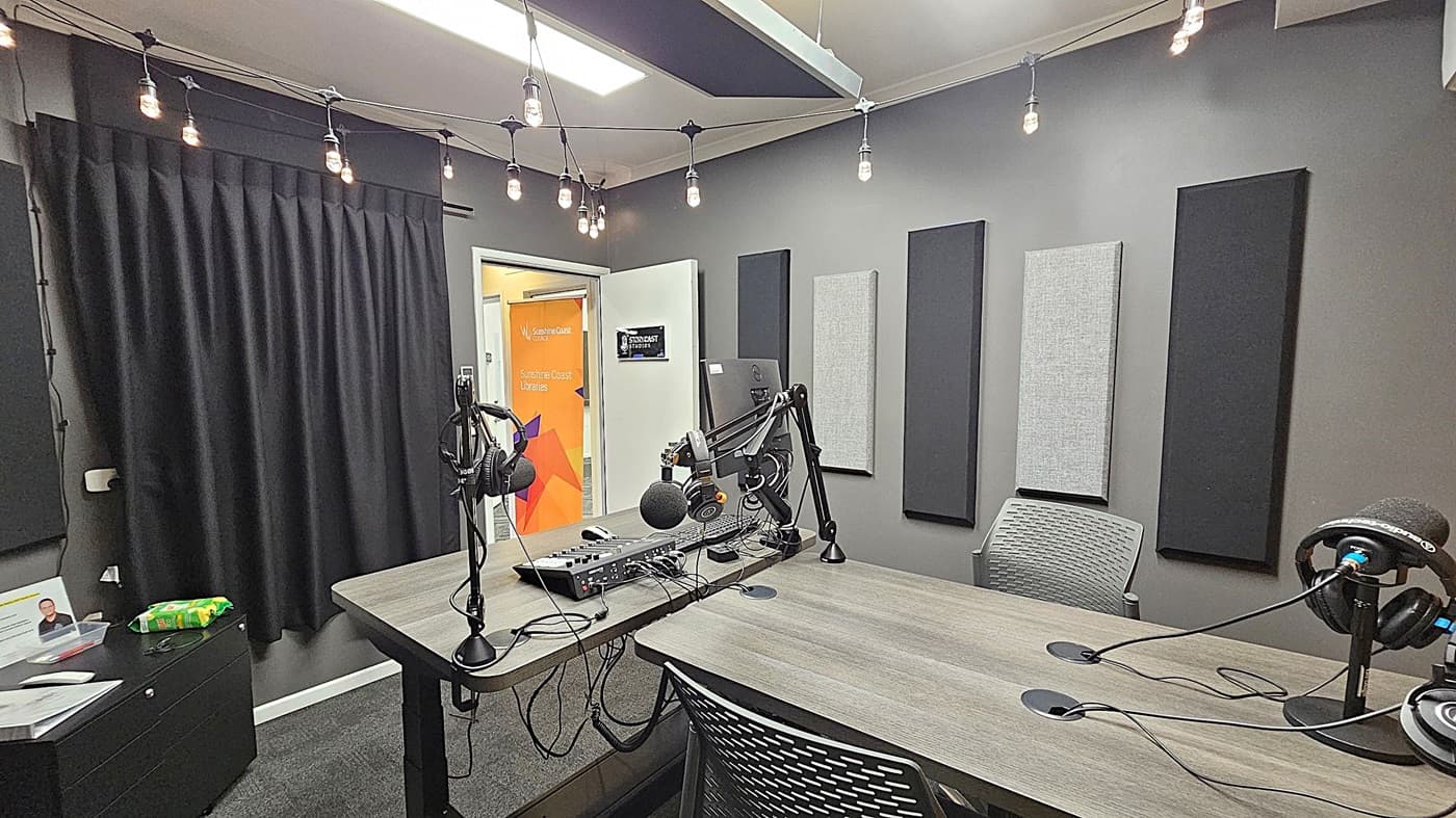 A podcast studio with a wooden table in the centre, equipped with microphones and headphones on adjustable arms. The walls are grey with acoustic panels. A string of exposed bulbs hangs from the ceiling. On the left, a black curtain covers part of the wall, and a small cabinet holds some items. In the background, a door is open, revealing a colourful sign with text. The floor is carpeted in dark grey.