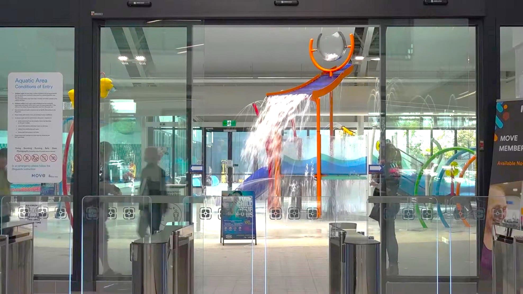 The entrance to the aquatic centre is through an automatic glass sliding door adorned with posters. On the left, a partially visible person stands behind frosted glass. Various posters and signs are affixed next to the door. To the right, an indoor playground with a water feature, including a slide, hoops, and splash areas, is visible. The ceiling and floor are both white. In the foreground is a turnstile gate.