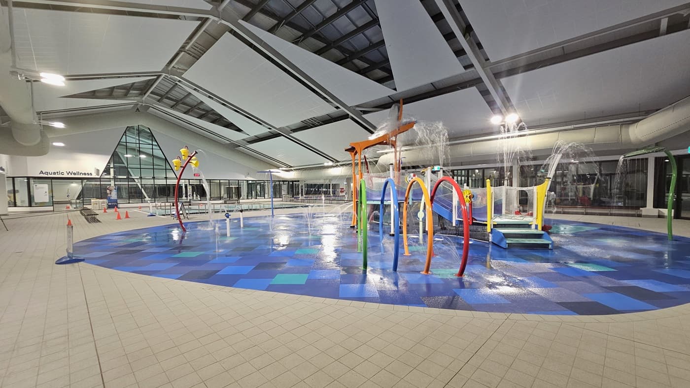 Indoor swimming facility with a children's water play area  featuring colourful hoops, sprinklers, and a slide. To the right, there are windows and a clear space leading to an adult pool barely visible in the background. The ceiling has exposed metal beams and skylights. The floor is tiled in blue and white with wet patches reflecting the overhead lights.