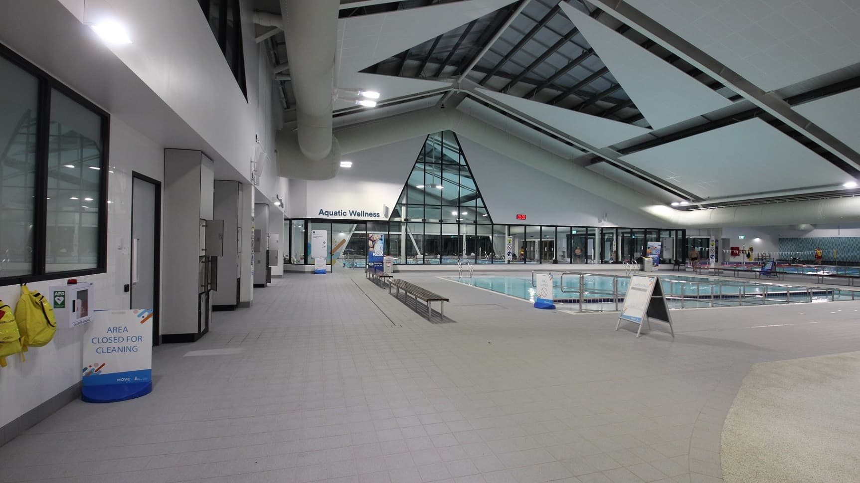 Inside the swimming facility, a sign reading "Aquatic Wellness" hangs above a large glass window, displaying a brightly-lit pool area. On the left, a corridor features several doors, signage, and a bench. To the right, the pool perimeter includes a ramp with handrails, lifeguard stands, and swimmers. Above, a white ceiling with recessed lighting illuminates the space. The glossy floor reflects the lighting.