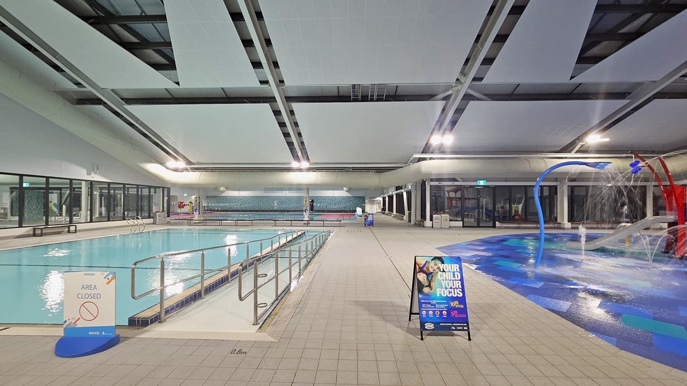 To the left is a pool with a ramp entrance, equipped with handrails. On the right is a children's play area featuring slides, sprayers, and colourful tiles. Free-standing banners are placed on the pool deck. The area is surrounded by glass. Above, a white ceiling with recessed lighting illuminates the space.