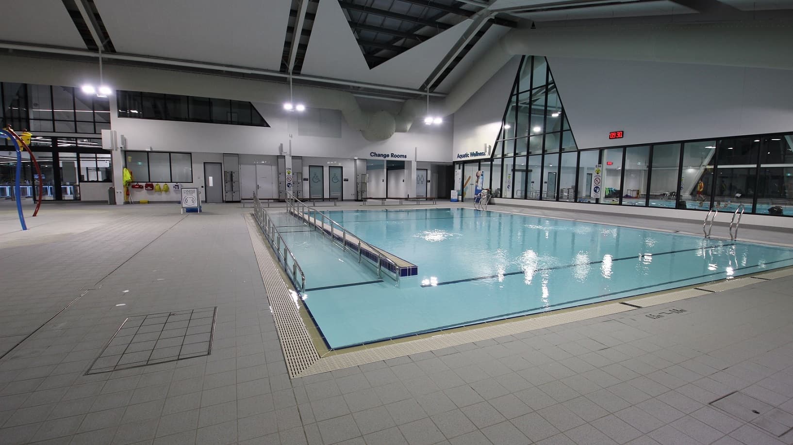 On the right, is an indoor swimming pool with calm water and marked lanes. The left side features the pool edge, diving blocks, and a lifeguard chair. The ceiling displays exposed beams and lights. A tiled floor surrounds the pool.