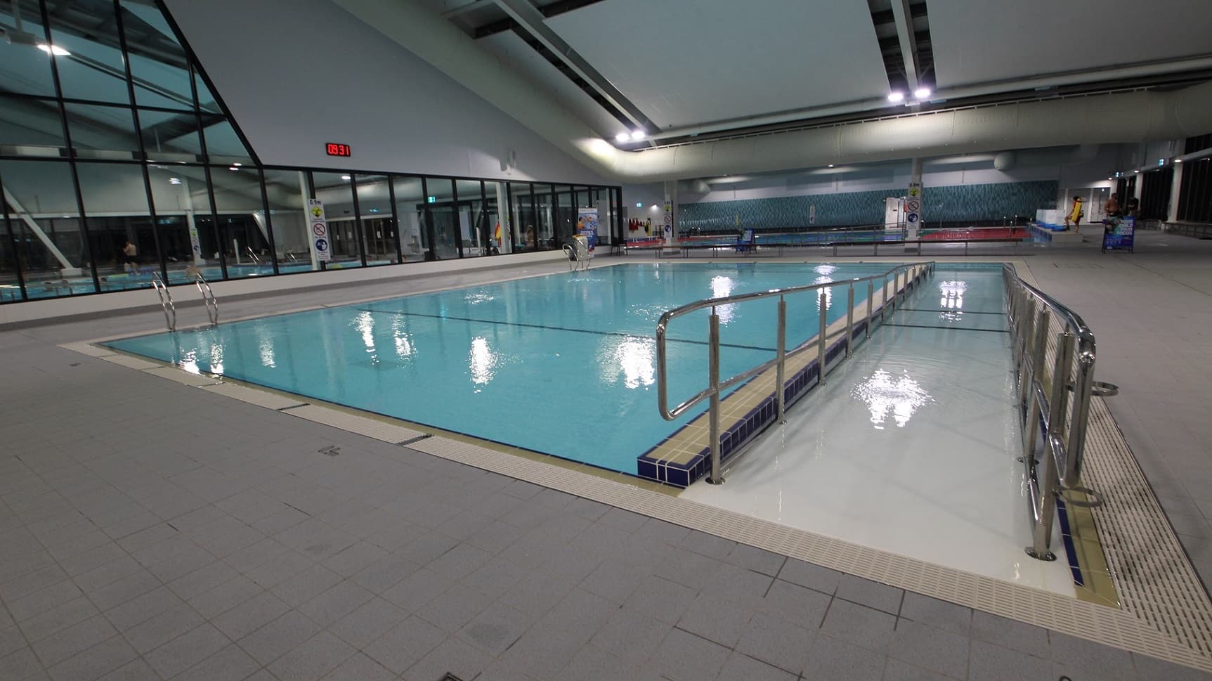 An indoor swimming pool equipped with a ramp and steps, both with handrails for entry. Bench chairs are arranged on the pool deck. Glass and dark turquoise chequered wall surrounds the space. The ceiling displays exposed beams, large air conditioning pipes, and recessed lights, while the floor is tiled in grey.