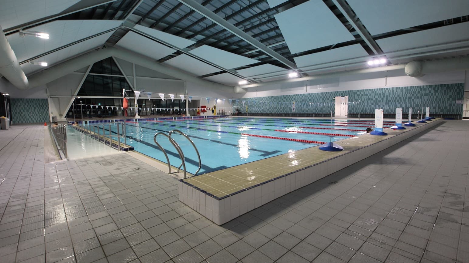 An indoor swimming pool is shown, equipped with a ramp and steps, both with handrails for entry. Floating ropes and triangular banners run across the water. Banners are placed on the raised pool deck. The walls feature a dark turquoise chequered design. A glass wall surrounds the space. The ceiling displays exposed beams, large air conditioning pipes, and recessed lights, while the floor is tiled in grey.