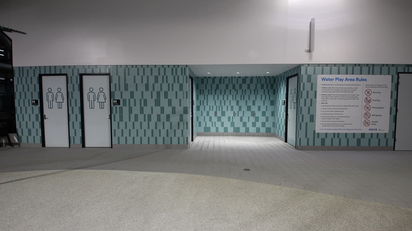 An indoor setting with a patterned light and dark turquoise chequered wall. To the left, two doors with male and female silhouettes signify restrooms. To the right, a large sign displays 'Water Play Area Rules' with bulleted text beneath. The ceiling is not visible. The speckled floor curves gently inwards towards the centre.