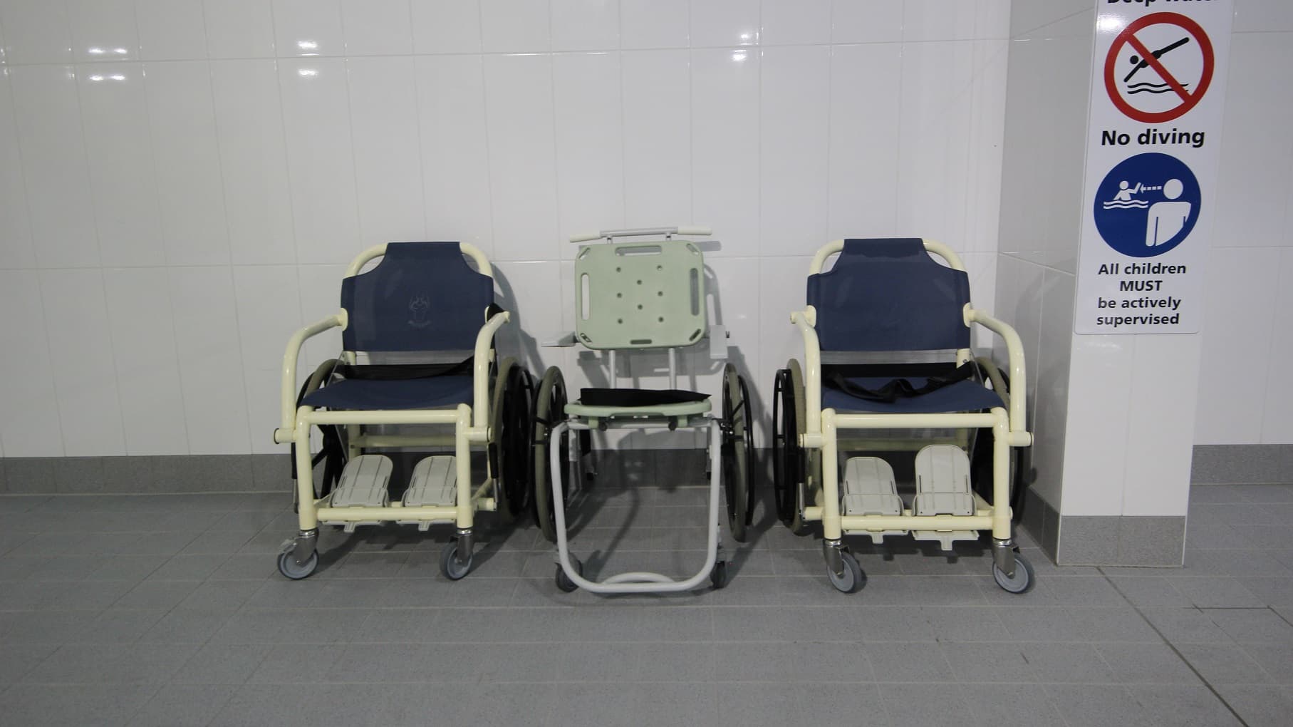 Three aquatic wheelchairs are positioned against a white tiled wall. The left and right wheelchairs have blue covers, while the one in the middle has no cover but features a light green backrest. The floor is tiled in grey. A signage poster is affixed to the column on the right.