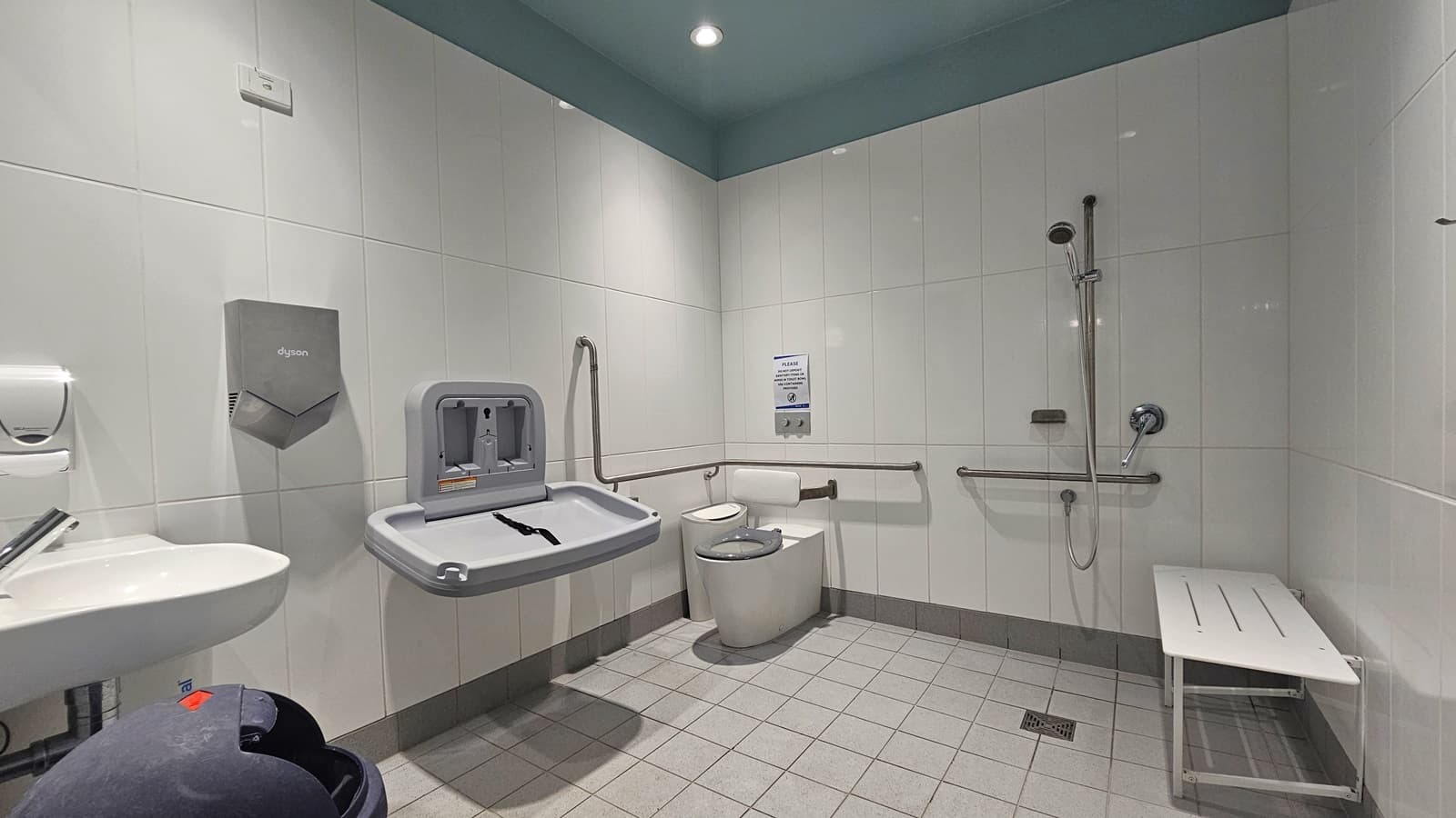 Within the accessible toilet, the toilet bowl with hand grabs is situated in the corner. Adjacent fixtures include a rubbish bin, change table, wash basin, liquid soap dispenser, and a hand dryer. To the right, a shower is mounted on the white wall alongside a bench chair. The floor is tiled in white, with a tortoise ceiling above.