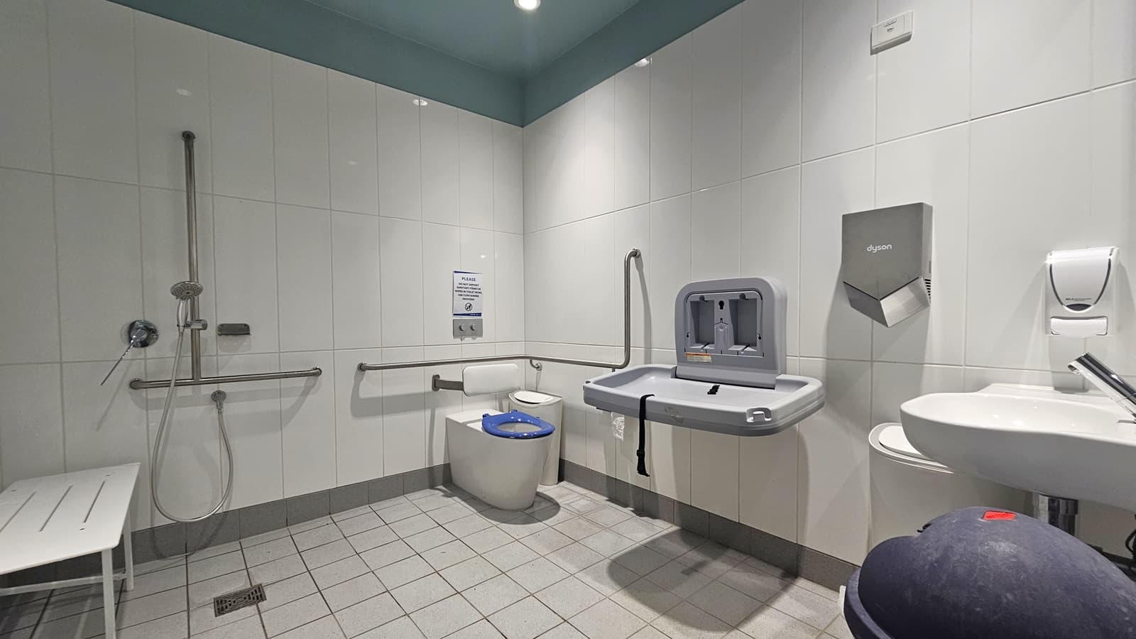 In the accessible toilet, the toilet bowl with hand grabs is in the corner. Nearby fixtures include a rubbish bin, change table, wash basin, liquid soap dispenser, and a hand dryer. On the left wall, a shower is mounted next to a bench chair. The floor is tiled in white, with a tortoise ceiling above.