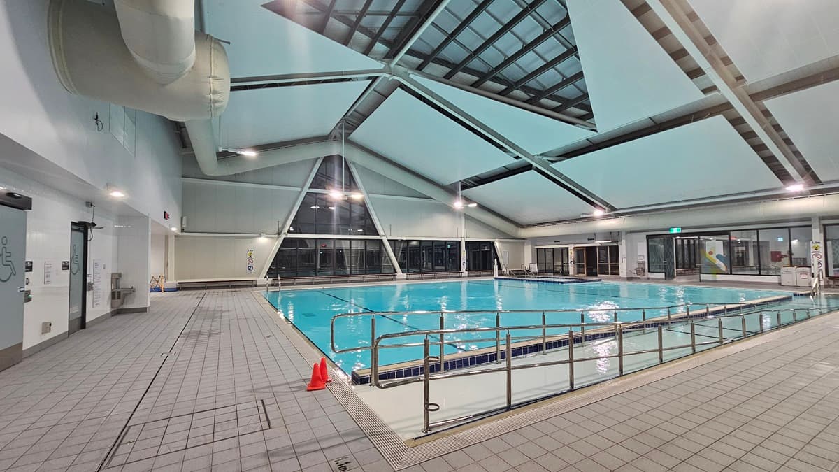 In an indoor pool area labeled 'Aquatic Wellness', a glass entrance is on the left with a blue poster detailing opening hours. On the right stands a banner stand. In the centre, a pool equipped with a ramp and handrails are partly visible, with safety cones on the deck. The ceiling features recessed lighting, exposed metal beams, skylights, and large air conditioning pipes. The floor is tiled grey.