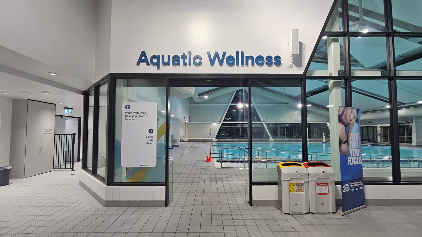 In an indoor pool area labelled 'Aquatic Wellness', a glass entrance is on the left with a blue poster detailing opening hours. On the right stands a banner stand. In the centre, a pool equipped with a ramp and handrails are partly visible, with safety cones on the deck. The ceiling features recessed lighting, exposed metal beams, skylights, and large air conditioning pipes. The floor is tiled grey.