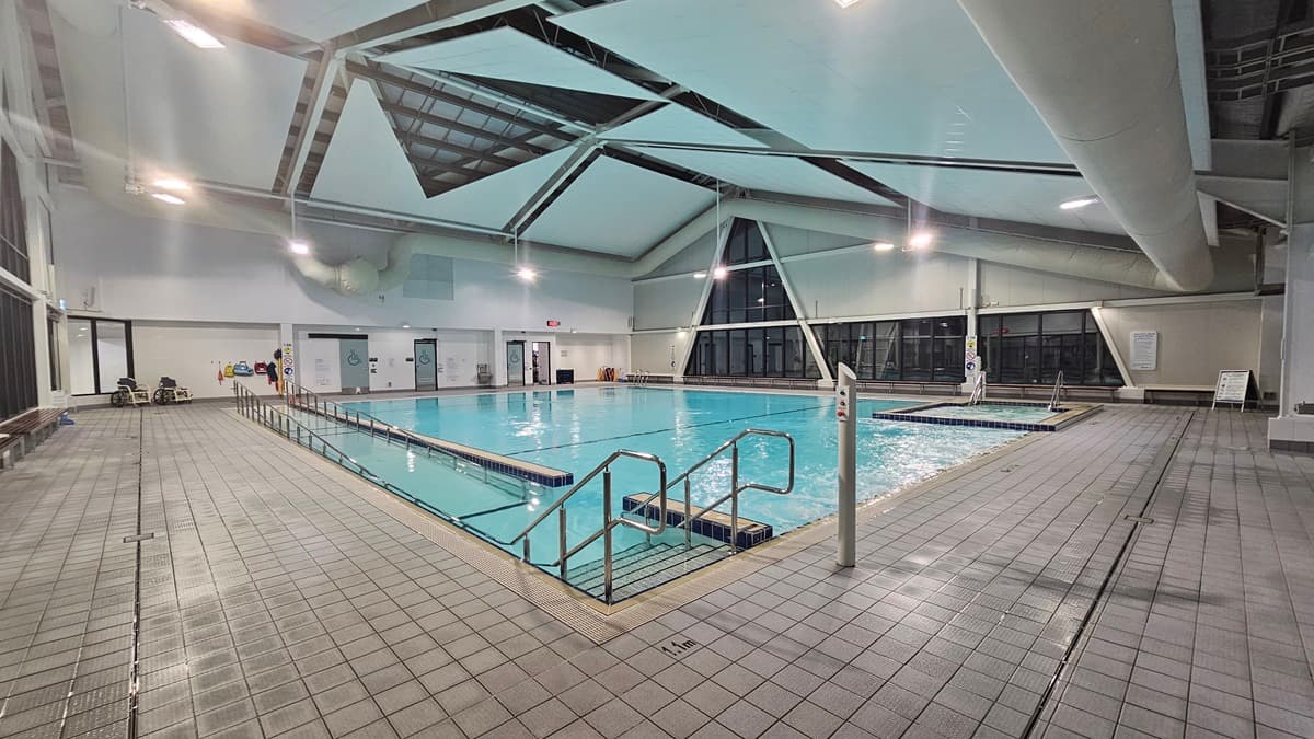 An indoor swimming pool is equipped with a ramp and handrails, as well as floating ropes. On the left, a wall features windows, doors leading to accessible change toilets and change rooms, and a drinking fountain. Bench chairs are positioned on the pool deck, along with two orange cones. The ceiling displays triangular beams and skylights, as well as large air conditioning pipes. The floor is tiled grey.
