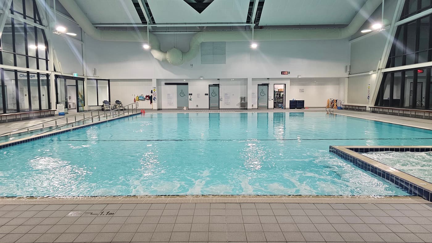 A closer view. An indoor swimming pool equipped with a ramp and stairs, both with handrails. Bench chairs are positioned against the glass wall on the left. At the far end, a door leading to accessible toilets is visible. Aquatic wheelchairs are arranged against the wall. Adjacent to the pool on the right is a spa. The ceiling features exposed beams and skylights. The tiled floor surrounds the pool.
