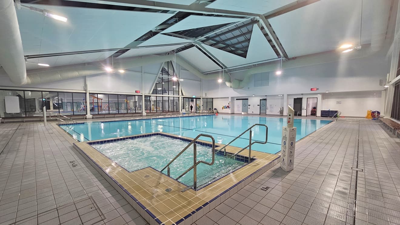 An indoor swimming pool equipped with a ramp and stairs, both with handrails. Next to the pool is a spa, also equipped with steps and handrails. Signage is visible on the walls and pool deck. Bench chairs are positioned against the glass wall on the left. The ceiling features exposed beams and skylights. The tiled floor surrounds the pool.