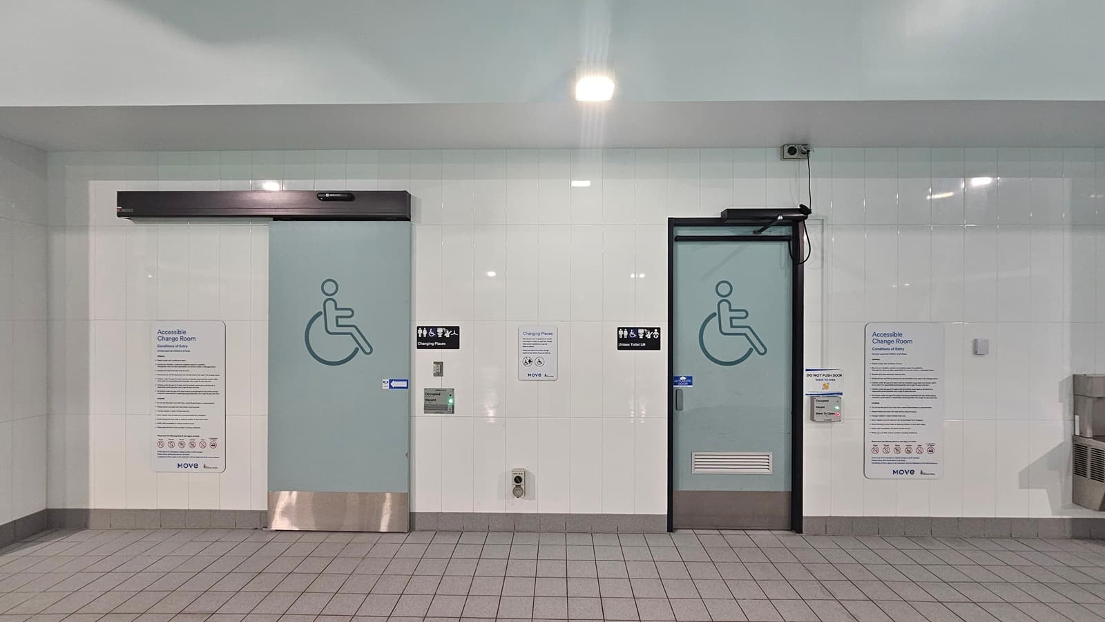 A restroom facility with tiled walls and floor. On the left, is a door with a wheelchair symbol and text; to its right, a similar door with tactile writing beneath the sign. Between doors, a panel with instructions in multiple languages. The ceiling is white with light fixtures and the floor has grey tiles.