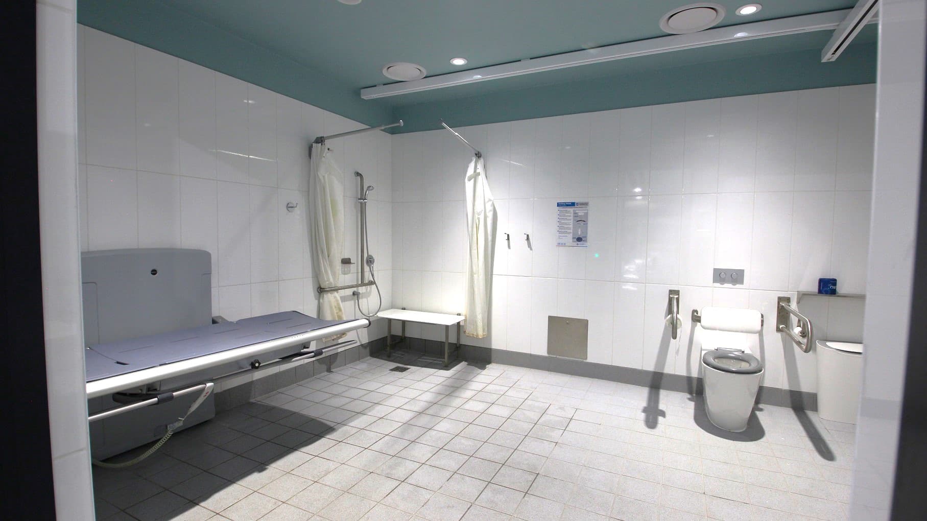 Inside the accessible bathroom. On the left, there's a foldable shower seat affixed to the wall and a white, horizontal, wall-mounted support bar above it. The right side features a toilet with a wall-mounted grab bar beside it and a bin in the corner. The ceiling has recessed lighting and there's a track for an overhead lift system. The bathroom includes tiled flooring. on the left, an adjustable adult-sized changing bench is visible, and information signs are seen on the wall.