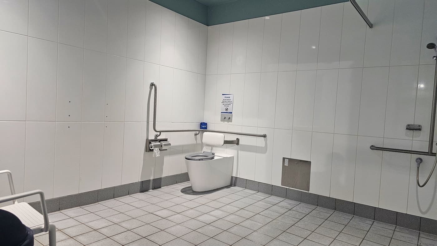 A spacious accessible toilet room with tiled walls and flooring. On the left, there's a fold-down grab rail and a wall-mounted assistance alarm cord. The right features a toilet with a grab rail above and a flush button to the side. A bin and toilet roll holder are adjacent to the toilet. The room includes a grey tiled floor and a white ceiling.