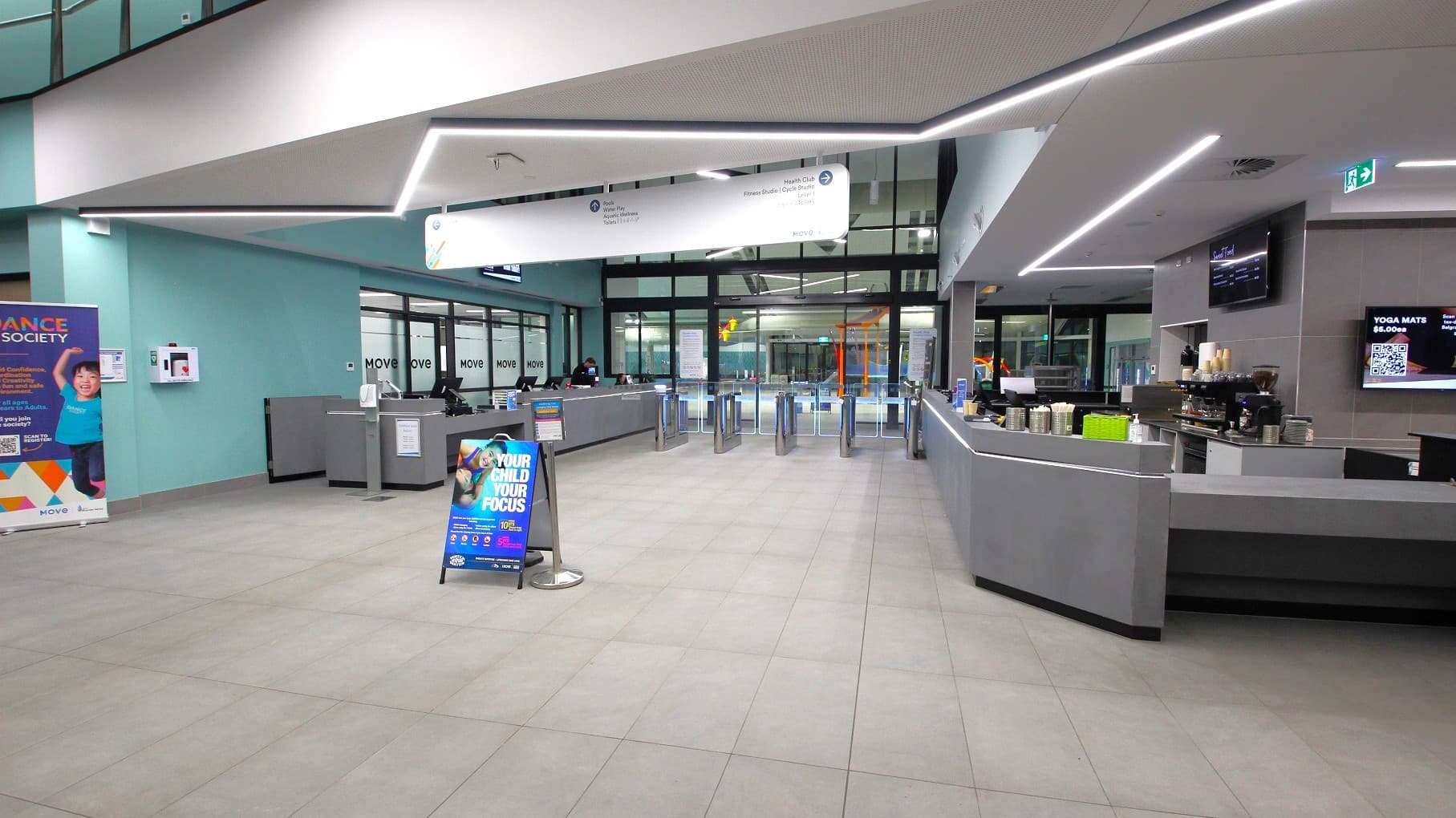 An indoor area has grey flooring and ceiling with recessed lighting and a large directional sign. To the left, there's a reception desk with a silver facade. On the right, a cafe counter holds multiple electronic cash registers and a menu display, with condiments arranged atop a cupboard. In the foreground, an open area features a banner and a couple of promotional stands. In the background, an entrance with turnstile gates is visible.