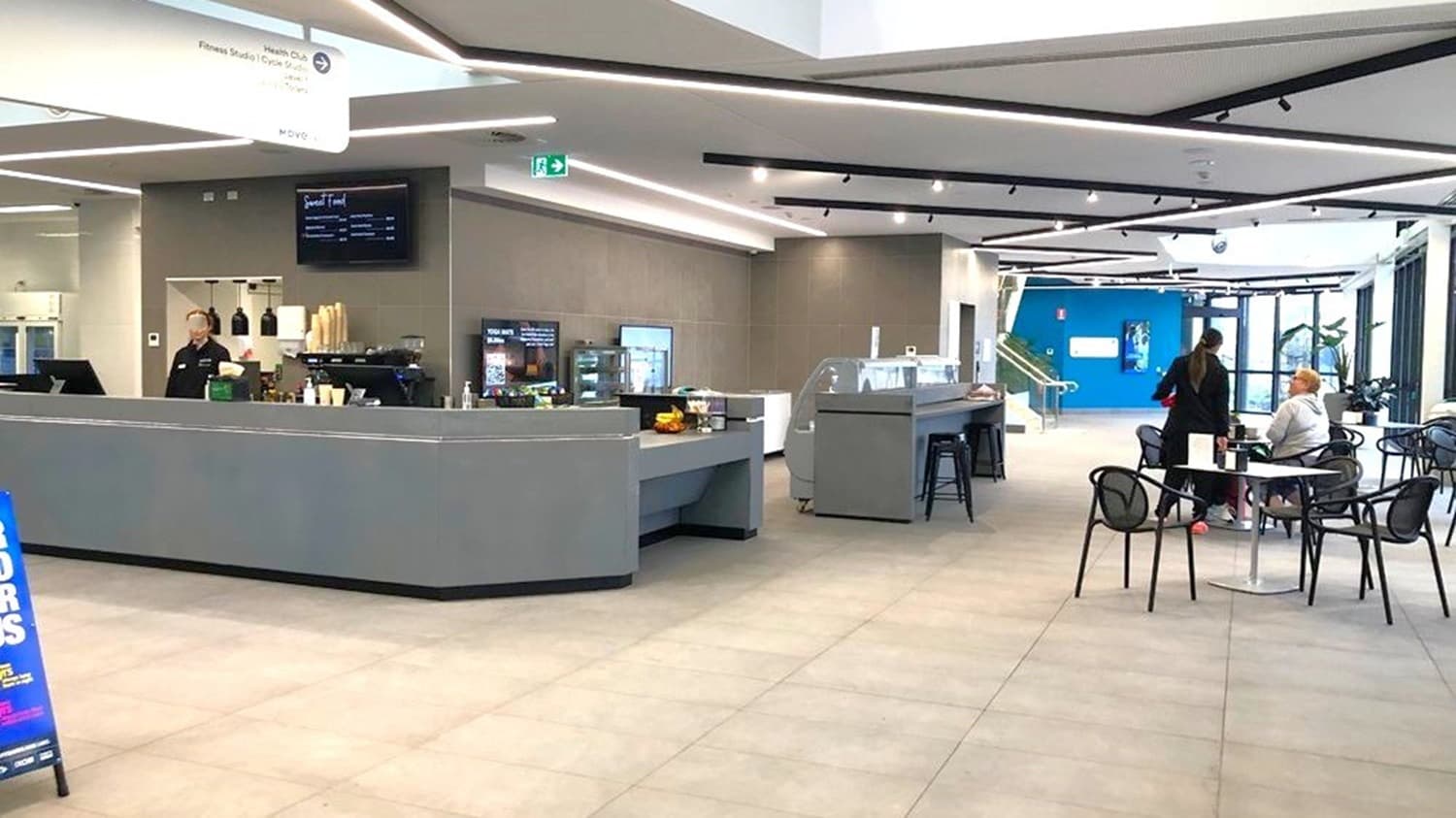 Indoors, grey flooring and ceiling are illuminated by recessed lighting, with a large directional sign. To the left, a cafe reception counter hosts multiple electronic cash registers and a menu display, with plastic cups and condiments atop a cupboard. On the right, plastic chairs and tables are arranged. In the foreground, promotional stands are featured. In the background, a blue wall and stairs with handrails are visible.