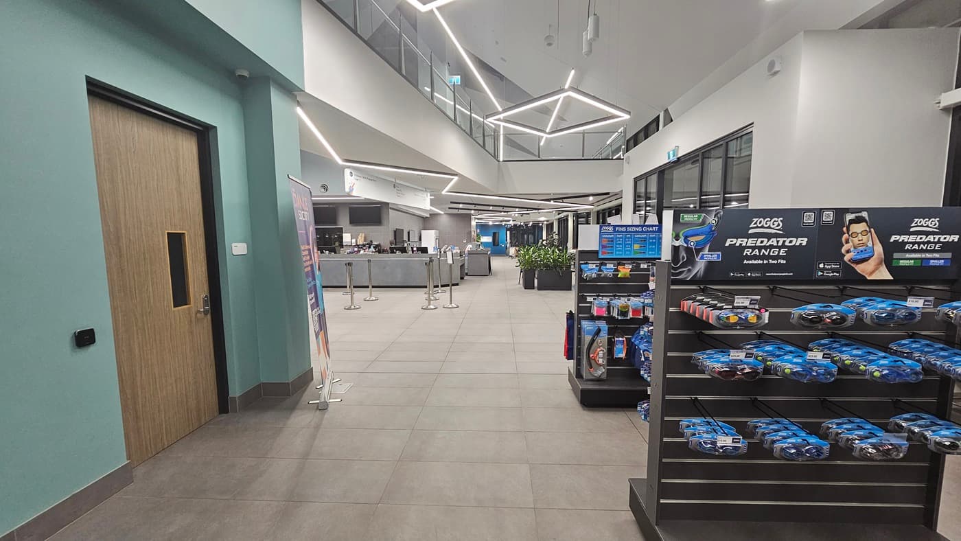 A small retail space with light grey flooring. On the left, is a closed wooden door with a digital keypad. The right features shelves with goggles and promotional stands. The ceiling has an intricate geometric design with recessed lighting. In the background, the cafe counter is visible. Queue bollards and planter boxes with indoor plants are positioned in front of the counter.
