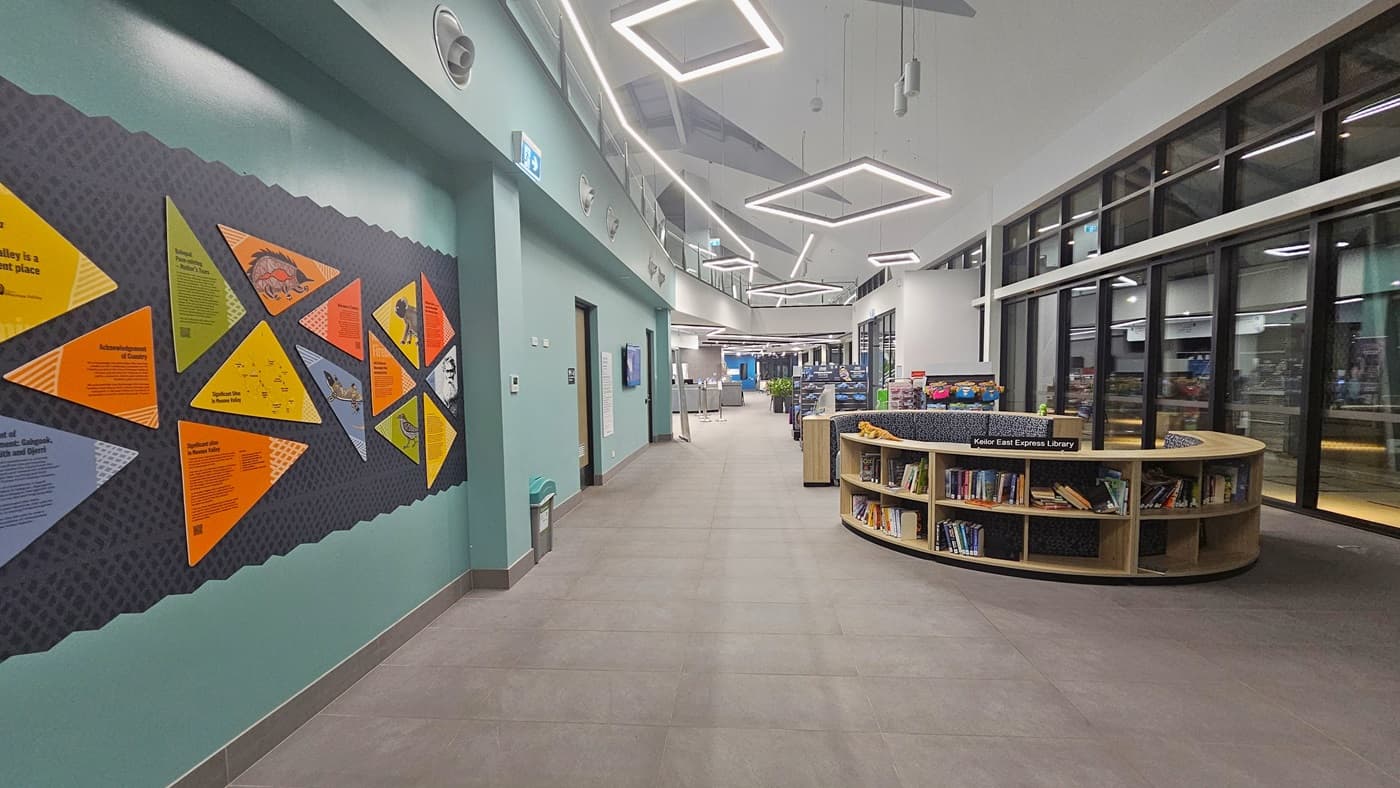 An indoor corridor. On the left, a wall with geometric-patterned decals and informational posters. The right side features a seating area with upholstered benches and a round bookshelf filled with books. Overhead, the ceiling is fitted with recessed and pendant lighting. The floor has grey tiling, guiding through the space.