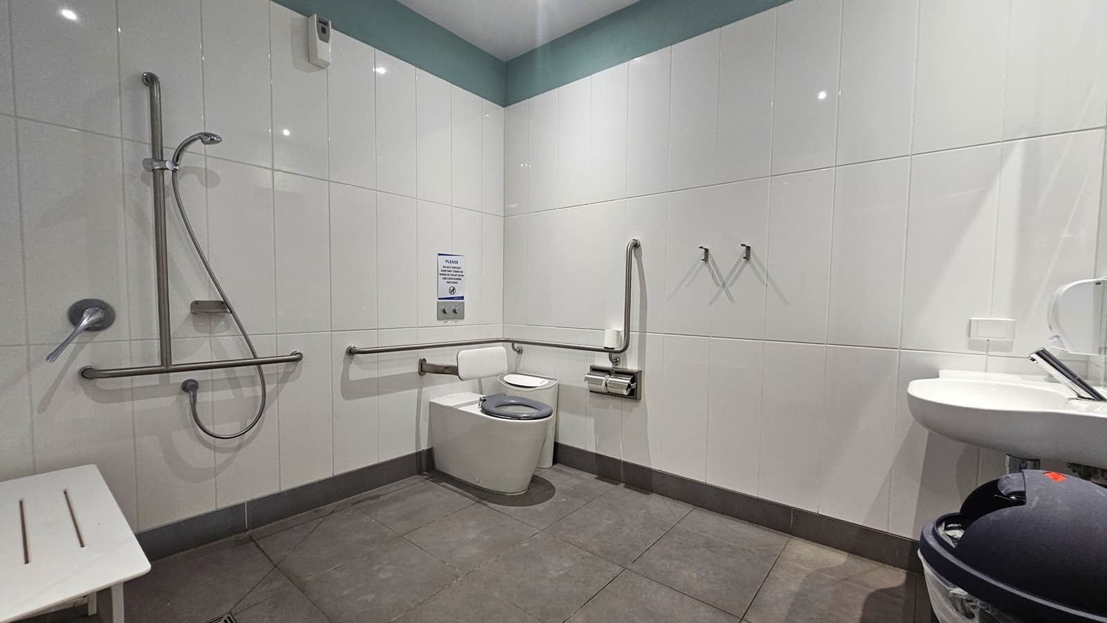 In the accessible bathroom, the toilet bowl with hand grabs is positioned in the corner. Beside it, a rubbish bin and a tissue dispenser are placed. To the left, a shower with hand grabs is affixed to the wall, with a bench chair beside it. On the right, a wash basin and a hand dryer are mounted to the wall, with a rubbish bin underneath.