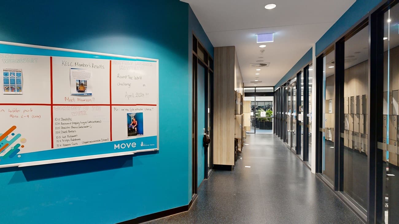An indoor corridor with blue walls. On the left, a bulletin board with various notices hangs next to a recessed, lit alcove with a door. On the right, glass doors with wooden frames lead to other rooms. Overhead, rectangular light fixtures are set into a grey ceiling. The floor is grey and the ceiling is white with recessed lights. The corridor extends into the background.