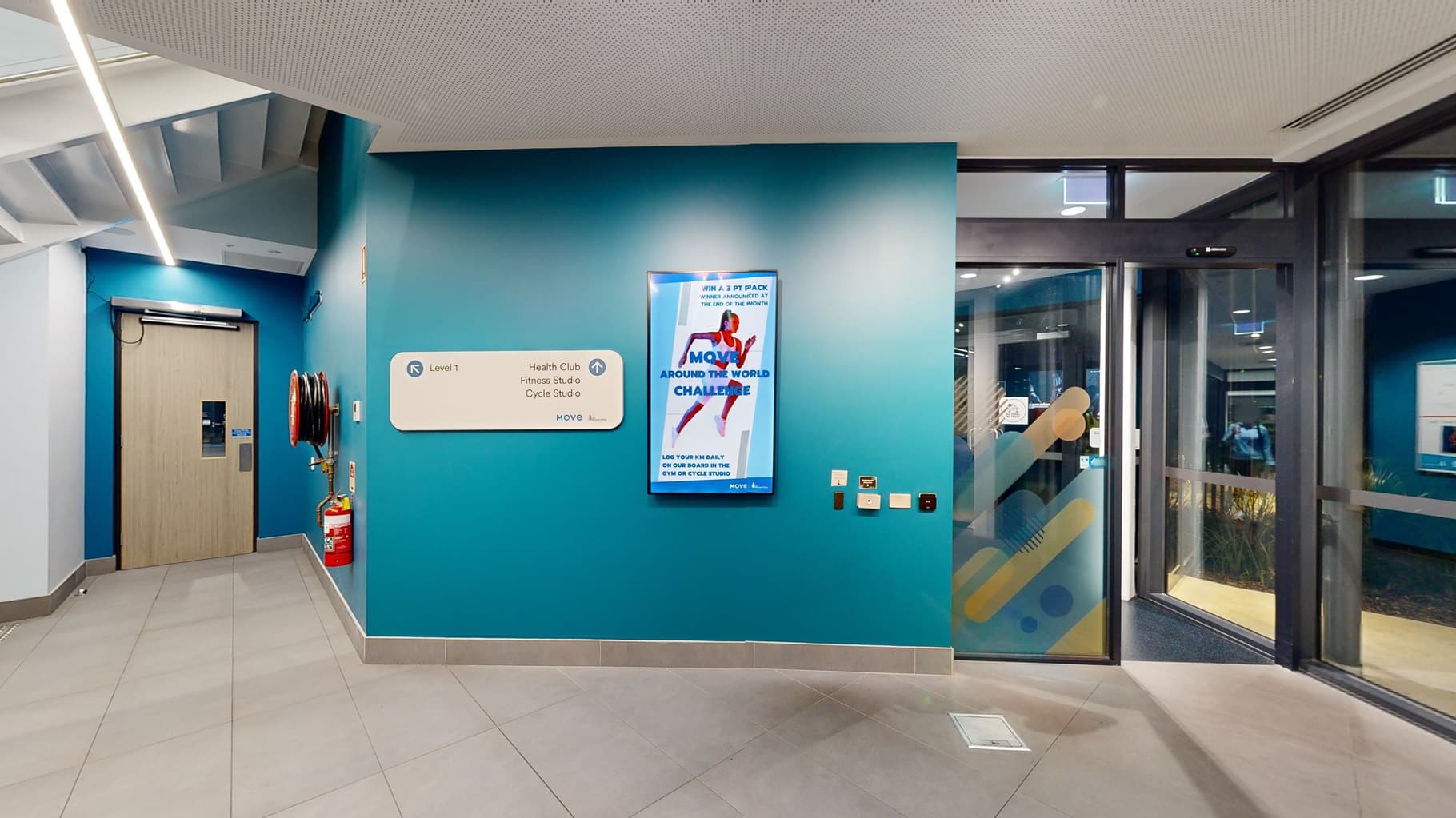 An interior space features a turquoise wall on the left, adorned with a poster and a white rectangular sign. Glass doors on the right offer a view into another room with yellow elements. On the left is a door with a timber front, accompanied by a fire extinguisher and a fire hose reel mounted to the wall. In the foreground, the grey floor transitions into carpet. The ceiling is white and features recessed lighting.
