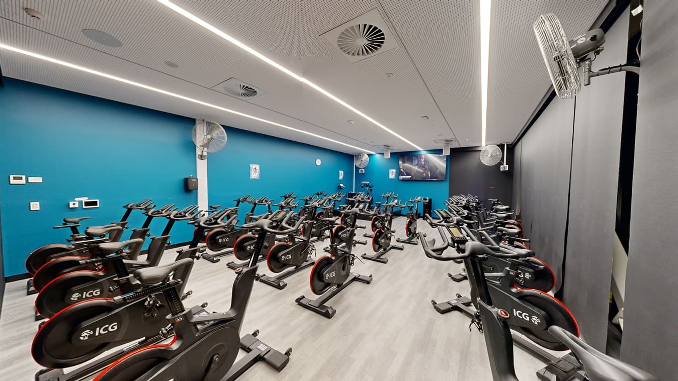 Interior of a gym with a row of ICG-branded stationary bikes, aligned towards a screen at the front. On the right, additional bikes are staggered behind. The room has blue walls with mirrors, wall fans, and a flat-screen TV mounted. The white ceiling features recessed lighting and air vents. The floor is covered in timber.