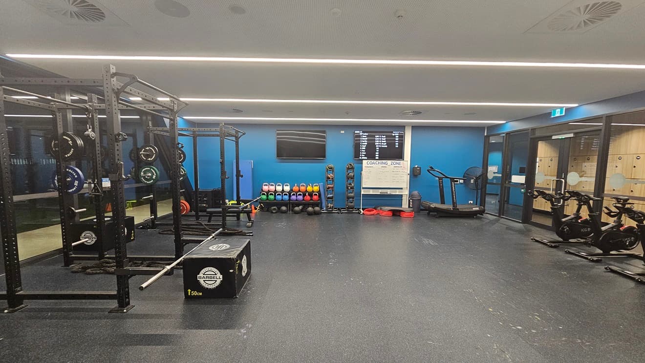 Inside the gym, Smith machines with weight plates and a barbell are on the left. On the right, a rowing machine, static bikes, and elliptical trainers are present. Kettlebells are arranged in a central rack, along with a wall-mounted flat-screen TV. Rubbish bins and a large fan are placed nearby. The ceiling has recessed strip lighting, and the floor has black rubber tiles. Both walls are painted blue, and lockers are visible in the background.