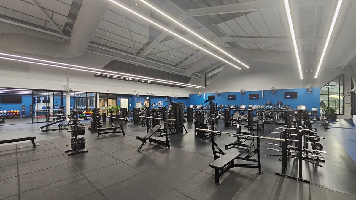 In an indoor gymnasium, weight benches, barbell racks, and dumbbell sets are in the foreground. Cardiovascular machines, including treadmills and ellipticals, are on the right. Kettlebells are displayed on a rack on the far left. The white ceiling has strip lighting, large air conditioning duct pipes, and insulation foams, while the floor is black rubber tiles. Flat-screen TVs and fans are mounted to the blue wall.