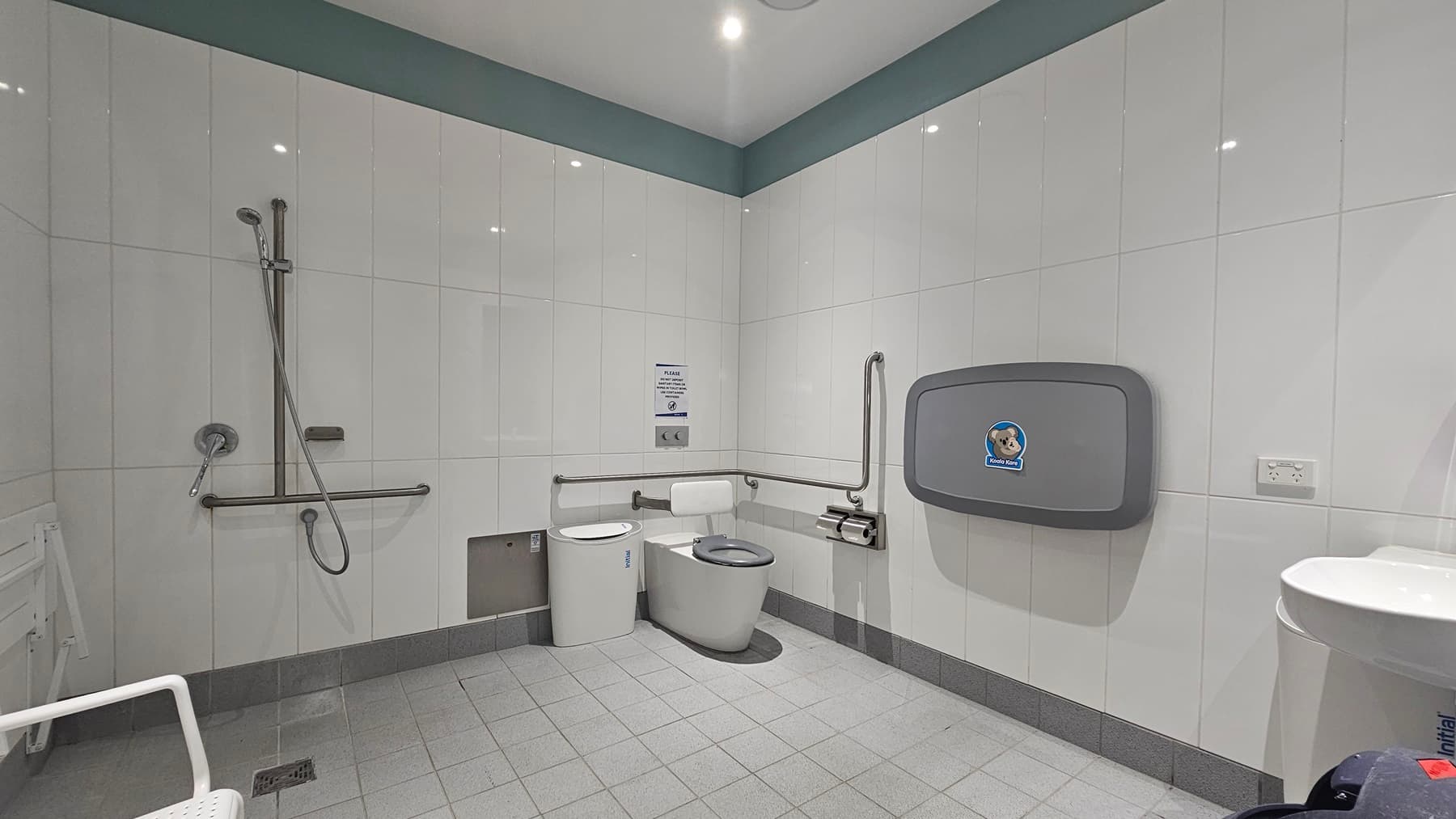 An accessible bathroom with white tiles. On the left, a wall-mounted shower with a stainless steel handrail. Next to it, a white toilet bowl with grab bars and an emergency cord. A white bench chair is mounted to the wall. To the right, a wash basin and a rubbish bin. A fold-down baby changing table is also on the right.