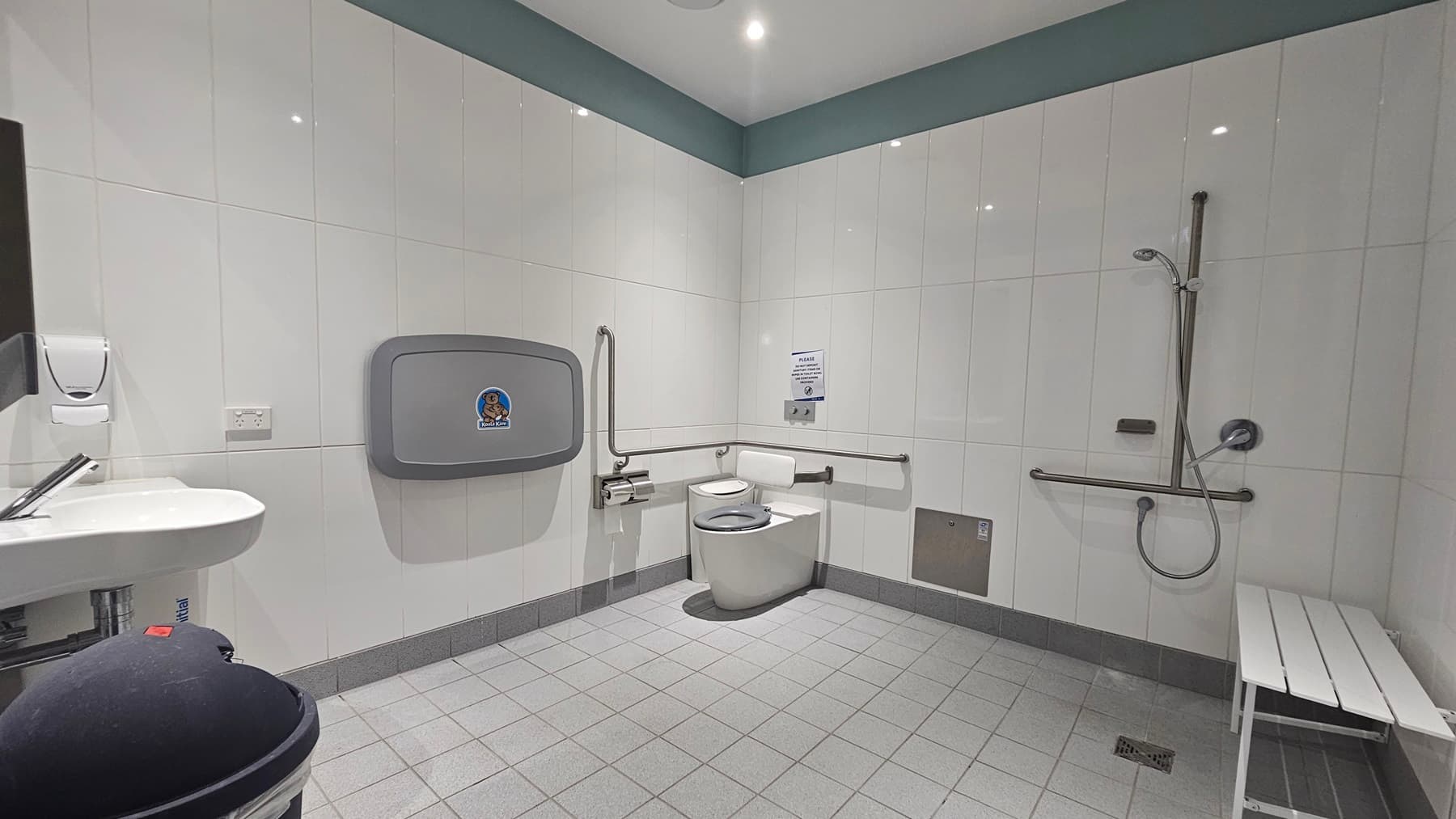 An accessible bathroom features light grey floor tiles. On the left, there is a wall-mounted hand basin, mirror, and soap dispenser. In the corner, a white toilet is equipped with grab bars, a folding support arm, and an emergency cord. To the right, there is a white bench and an accessible shower with a handrail. The ceiling and walls are white.