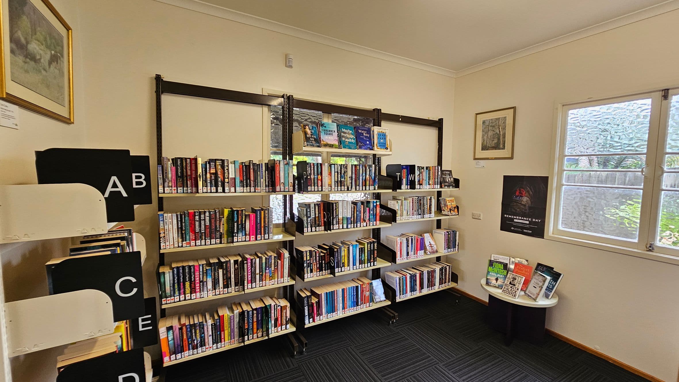 Bookshelves filled with books are against a cream-coloured wall. On the left, there are labelled sections with letters A to E. In the middle, three shelves hold books with dividers marked with letters. On the right, a small round table displays more books. Above it, a framed picture and a Remembrance Day poster are on the wall. A window with frosted glass is next to the table. The floor is carpeted with a dark pattern.