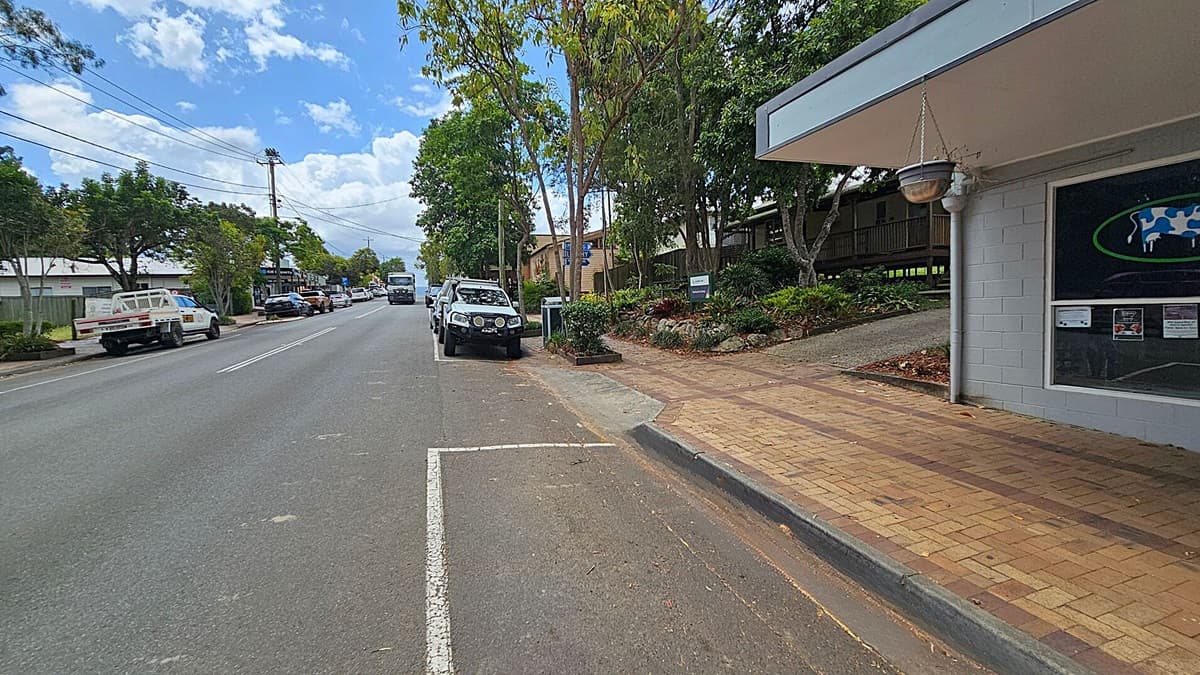 A street scene with a paved footpath on the right, bordered by a grey building with a hanging pot and a window displaying a cow graphic. Several cars are parked along the road, including a white vehicle in the foreground. Trees line the street, providing shade. On the left, more vehicles are parked, and a few buildings are visible in the distance. The road is marked with white lines, and the sky is partly cloudy.