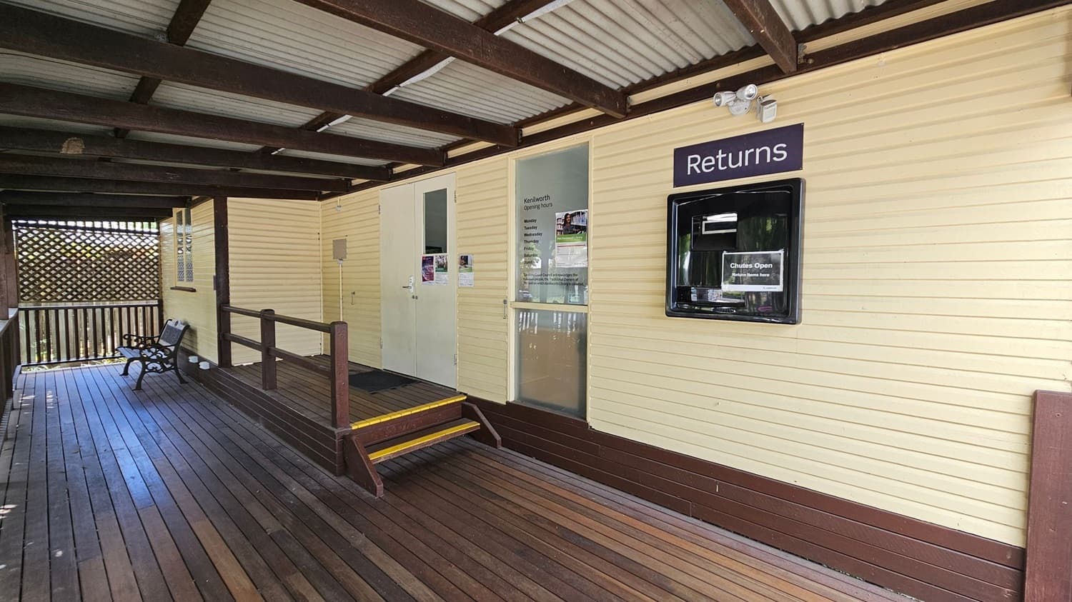 A wooden deck with a bench on the left side, leading to a building with cream-coloured walls. The building has a "Returns" slot on the right, and a glass door with opening hours listed. A small set of steps with yellow edges leads up to the door. The ceiling is made of corrugated metal with wooden beams. A lattice screen is visible in the background on the left.
