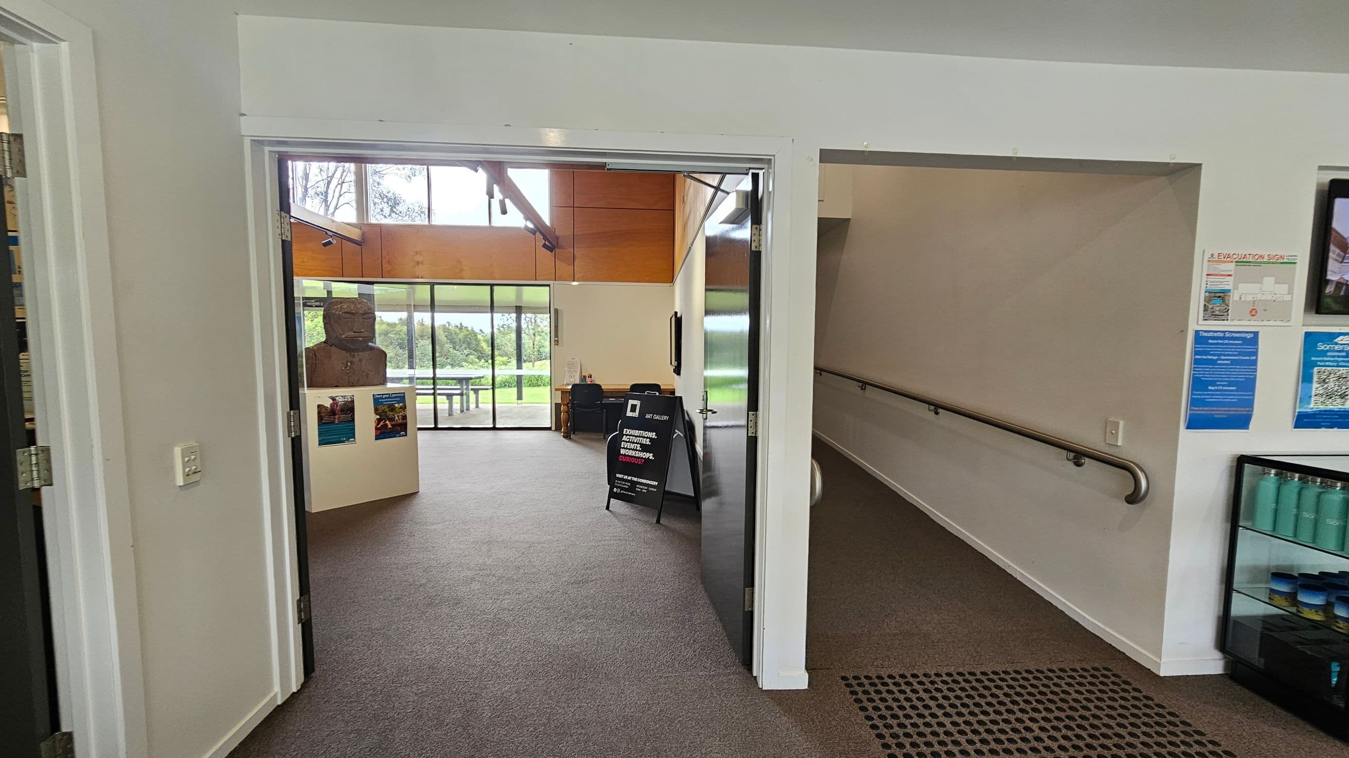 On the left, a doorway leads into a room with a large sculpture on a pedestal. The room has a carpeted floor and large windows showing greenery outside. In the middle, a black signboard stands near the entrance. On the right, a hallway with a handrail and informational posters on the wall. A display case with blue bottles is visible. The ceiling is white, and the floor is carpeted.