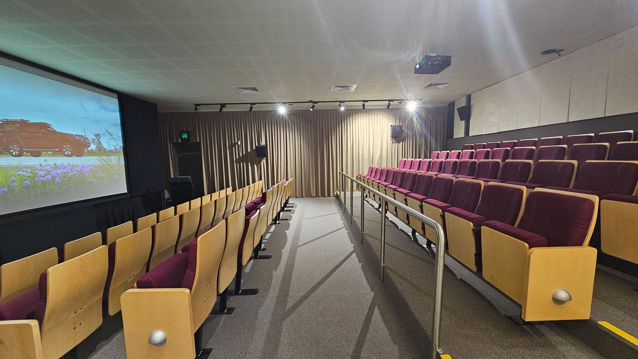 A small cinema or lecture room with tiered seating. On the left, there is a large screen displaying an image of a red vehicle on a road with flowers in the foreground. The seats are upholstered in maroon fabric with light wooden frames. A central aisle with a metal handrail divides the seating. The floor is carpeted in a grey and green pattern. The ceiling is fitted with recessed lighting and a projector. Beige curtains cover the back wall.
