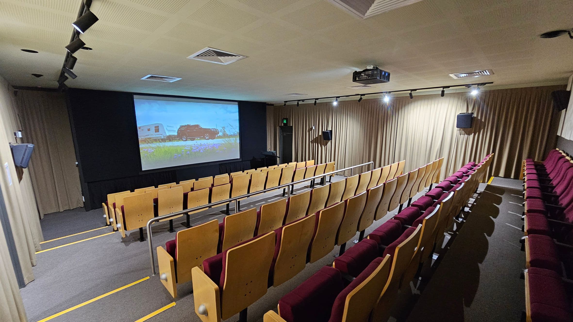 A small cinema or lecture room with tiered seating. The seats have wooden backs and red cushions. A large screen at the front displays an image of a car towing a caravan. The walls are covered with beige curtains, and there are speakers mounted on them. The ceiling is fitted with a projector and several spotlights. The floor is carpeted in grey with yellow safety lines on the steps.