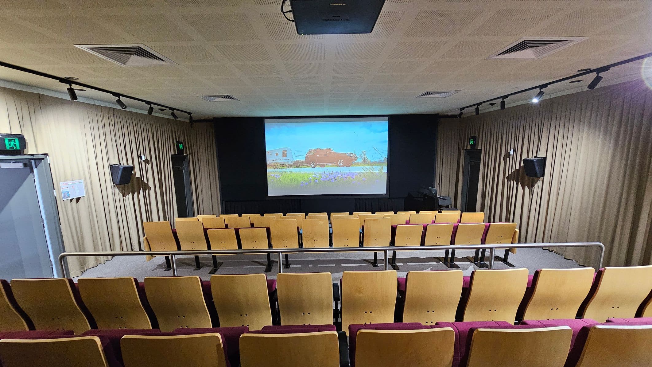 A small auditorium with rows of wooden seats with maroon cushions facing a large screen at the front. The screen displays an image of a red vehicle and a trailer in a field. Beige curtains line the walls, and there are speakers mounted on them. The ceiling is fitted with recessed lighting and air vents. Emergency exit signs are visible above doors on both sides. The floor is carpeted, and a metal railing separates the seating area from the aisle.