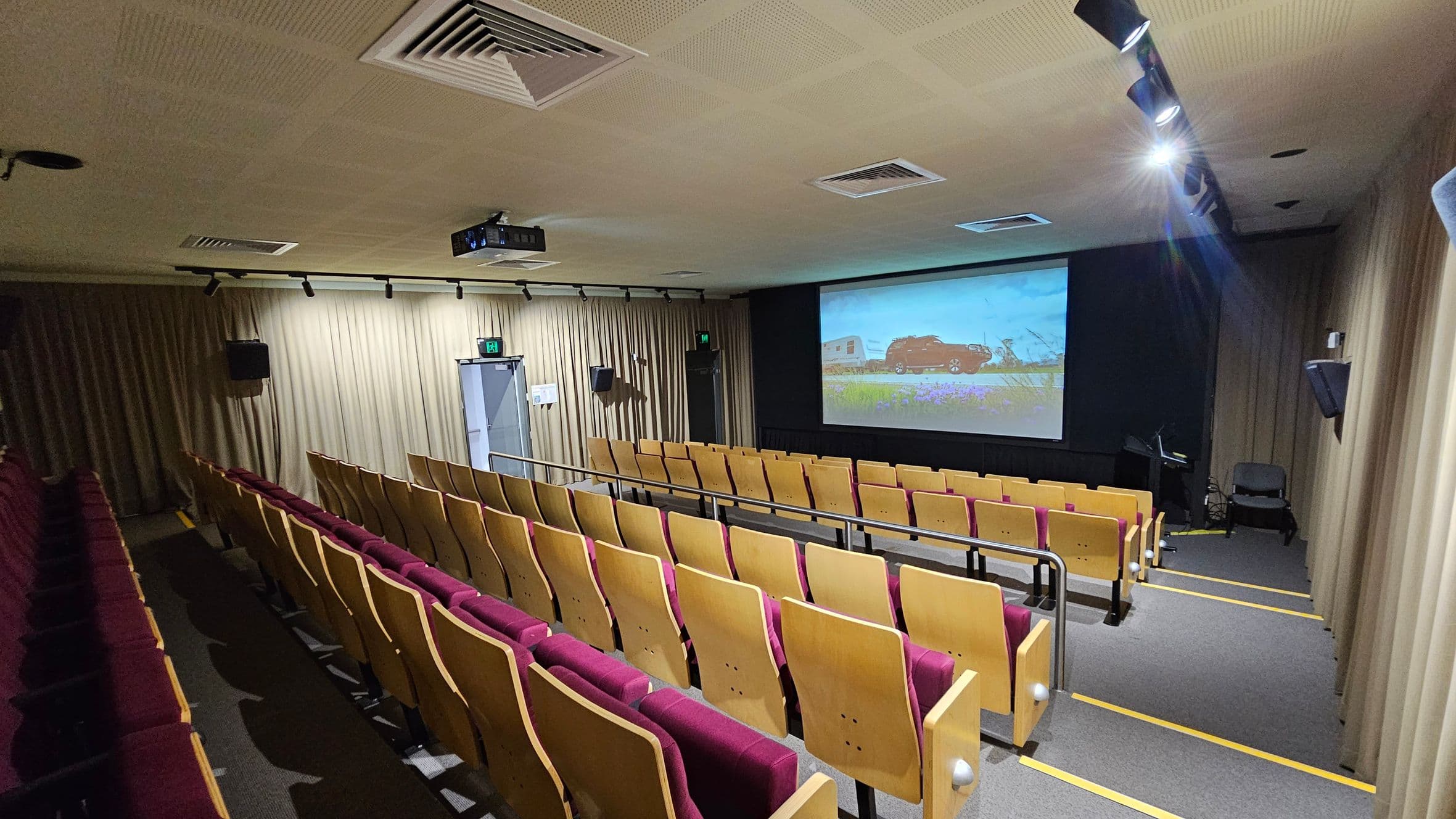 A small auditorium with rows of wooden seats featuring maroon cushions. The seating is arranged in tiers, facing a large screen displaying an image of a car and caravan in a field. The walls are lined with beige curtains, and there are speakers mounted on them. The ceiling has recessed lighting and air vents, with a projector mounted centrally. An exit door is visible on the left side of the room. The floor is carpeted in grey with yellow step markings.