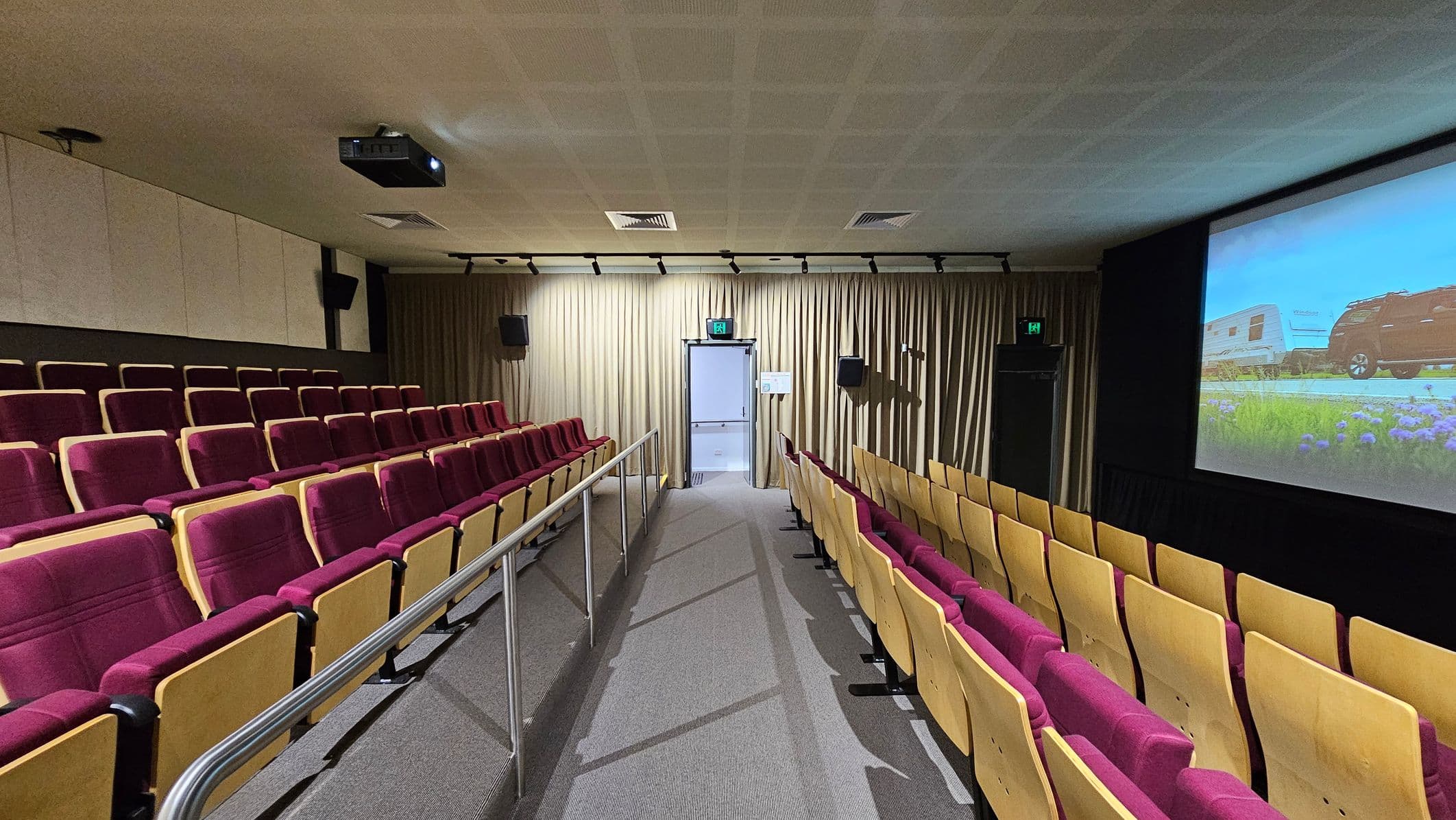 A small cinema room with rows of red and yellow seats on both sides of a central aisle. The floor is carpeted in grey, and a metal railing runs along the aisle. At the front, a large screen displays an image of a vehicle and trailer. The ceiling is fitted with a projector and ventilation panels. Beige curtains cover the back wall, where an exit door with an illuminated sign is located. Black speakers are mounted on the walls.