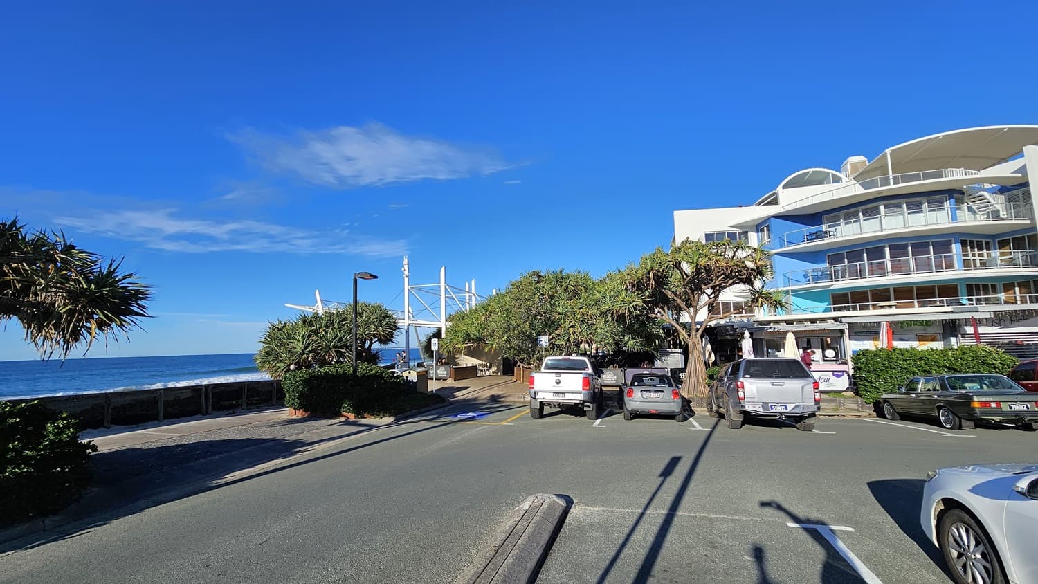 On the left, there is a coastal walkway lined with bushes and a tree, leading to the ocean. In the middle, there is a pathway with people walking, flanked by parked cars and trees. On the right, there is a multi-storey building with balconies and glass railings, adjacent to more parked cars. The foreground shows a carpark with multiple vehicles parked. The floor is a mix of asphalt and concrete.