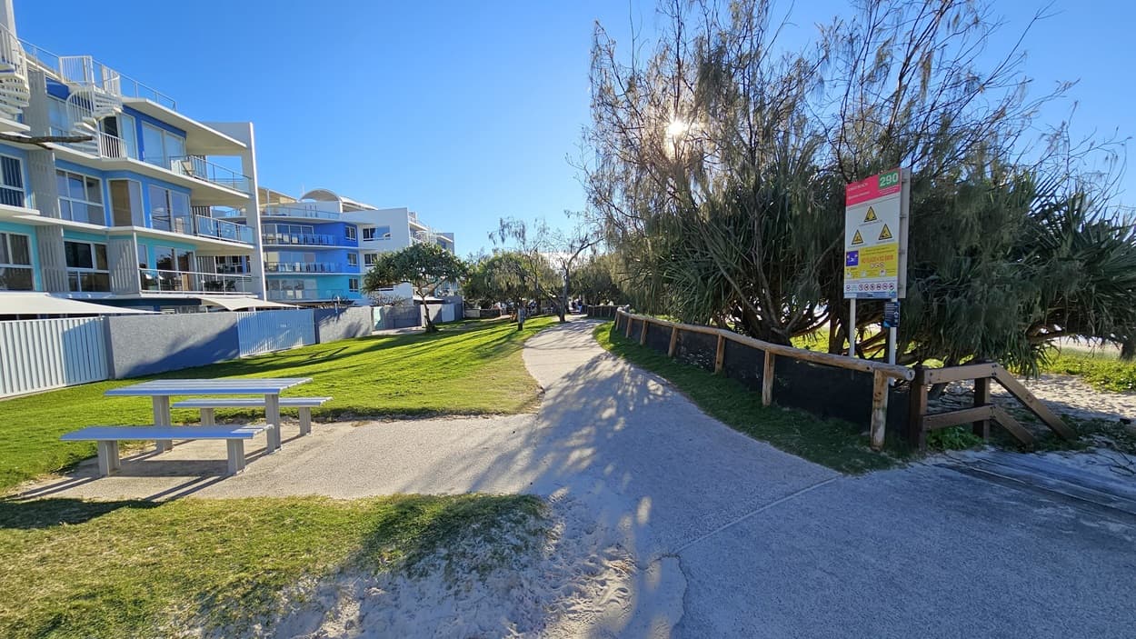 On the left, there is a grey picnic table on a grassy area, with a multi-storey building featuring balconies and glass railings behind it. In the middle, there is a winding concrete pathway bordered by grass, leading towards trees and more buildings in the background. On the right, there is a wooden fence with a large sign displaying warnings and information, next to dense bushes and trees. The floor is a mix of concrete and sand and there is a picnic table visible on the left.